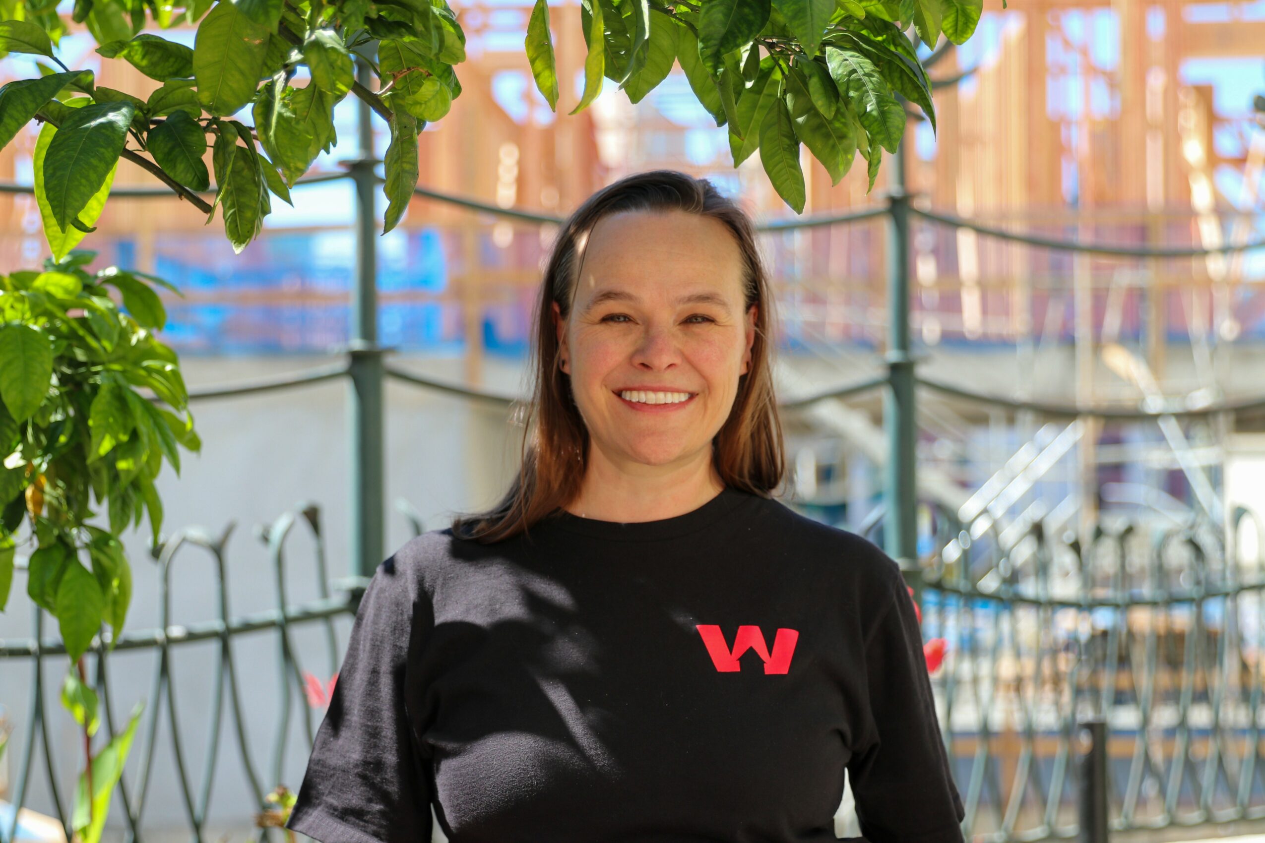 Smiling woman standing outdoors with trees and a marina in the background.