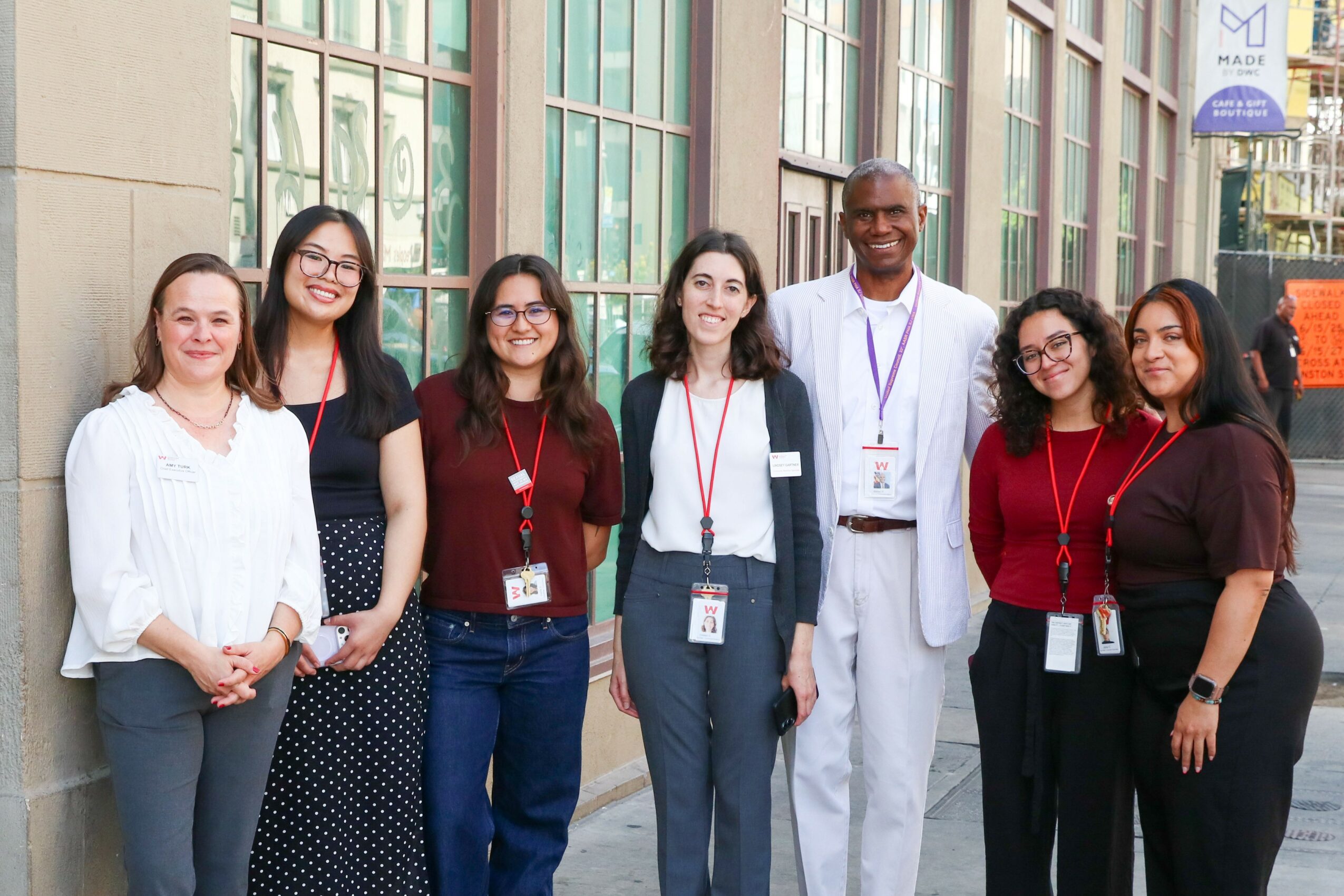 Group of seven diverse people standing outside a building, smiling, wearing conference badges, dressed in casual and professional attire.