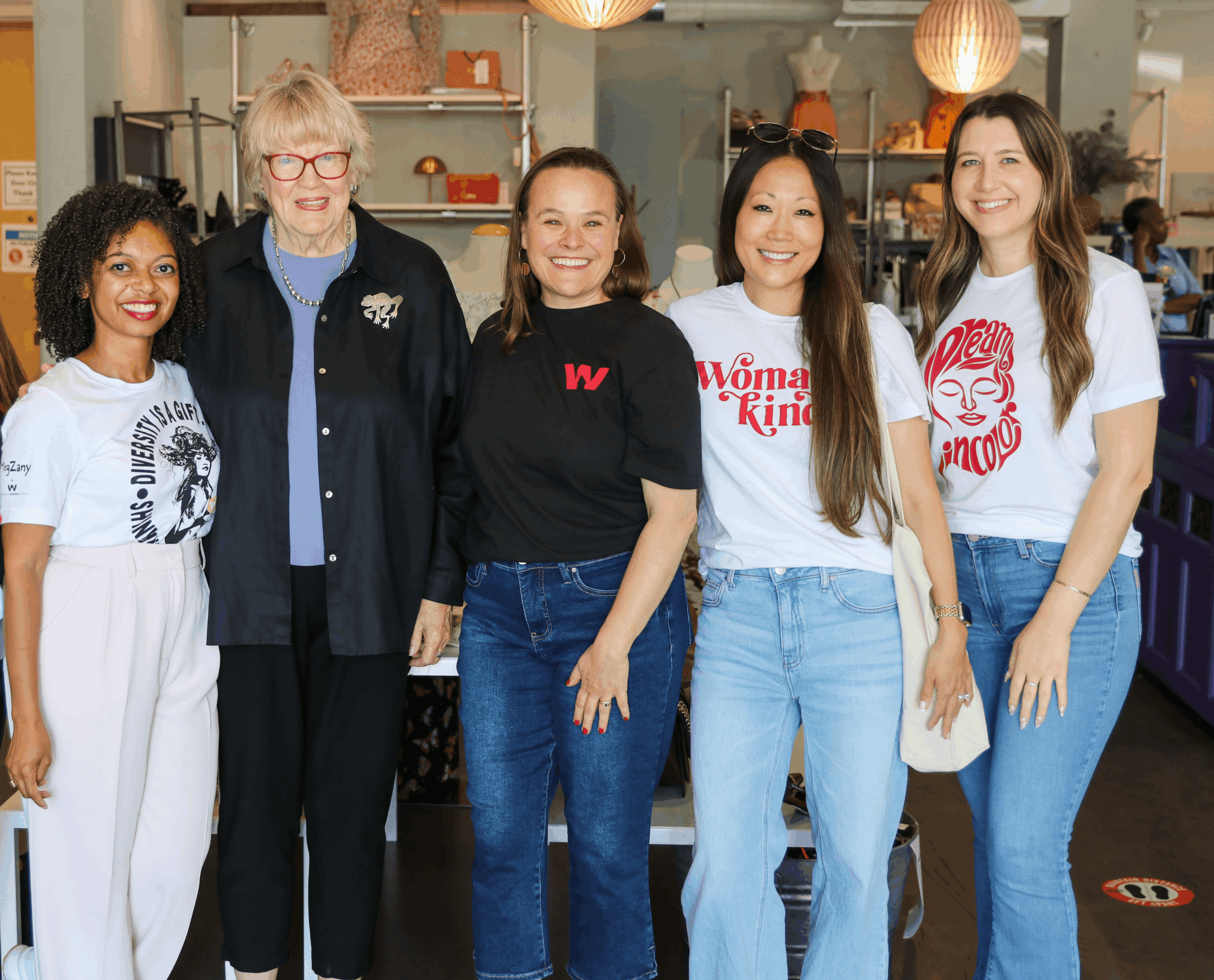Group of five women standing indoors, smiling, with shelves and lighting in background.