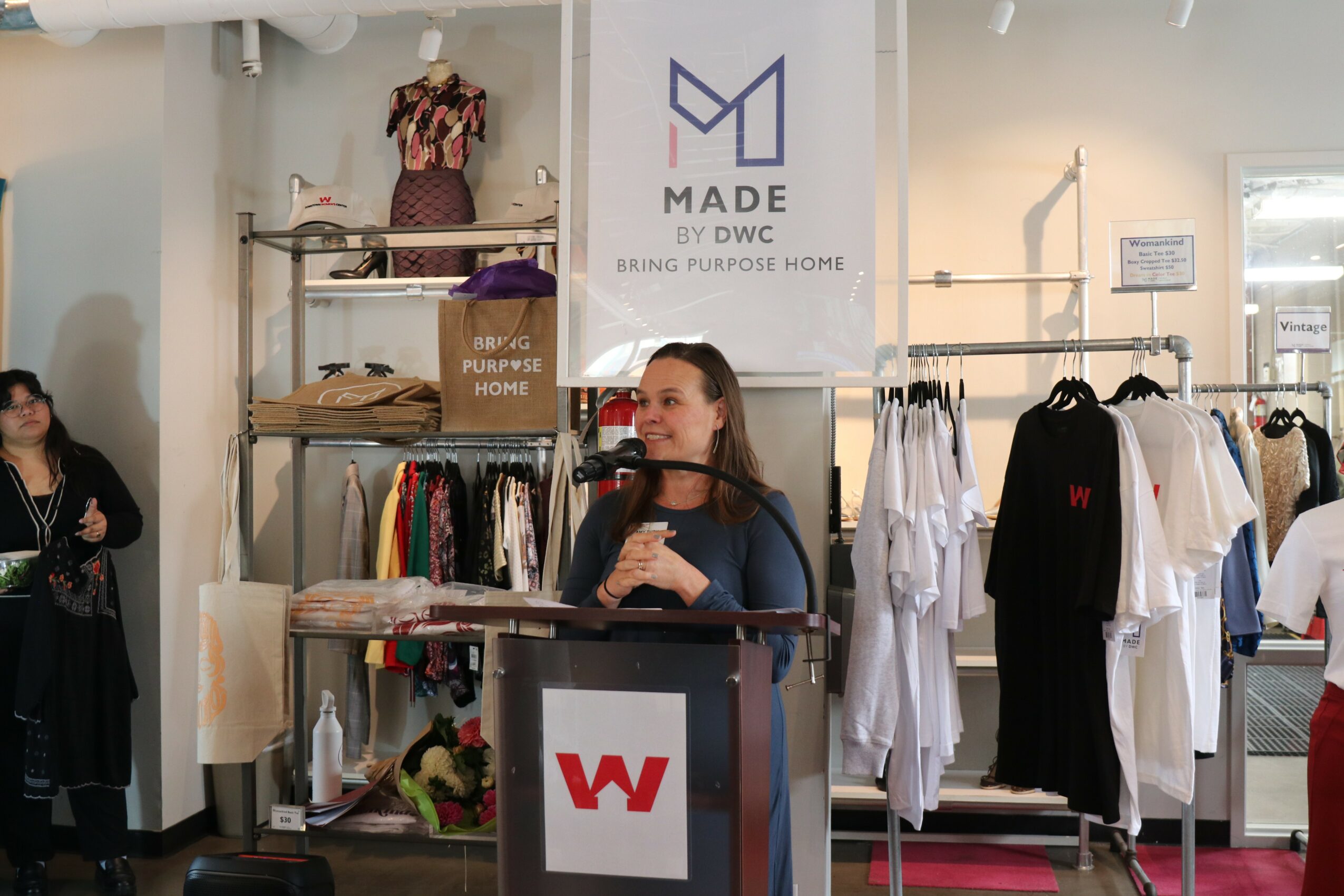 Woman speaking at a booth with clothing displays and a banner in the background, inside a retail space.