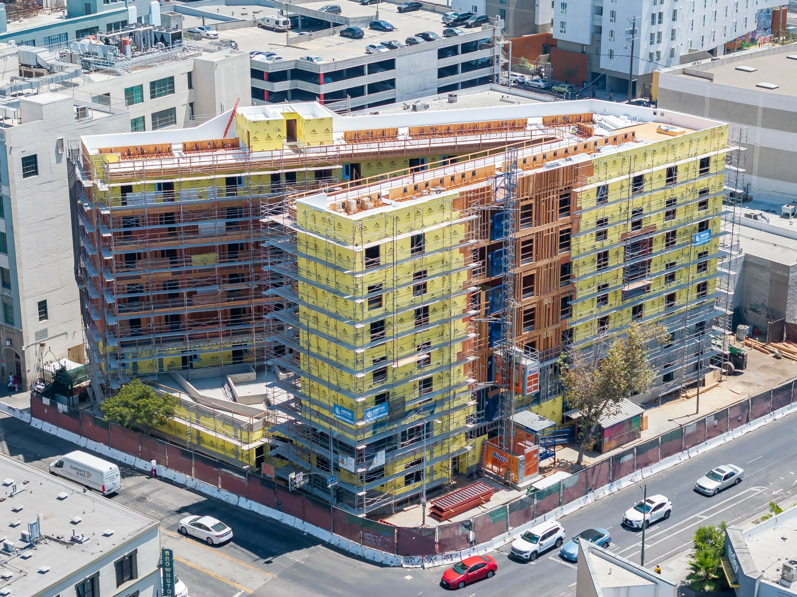 Construction site of a multi-story building with scaffolding, surrounded by city streets and parked cars.
