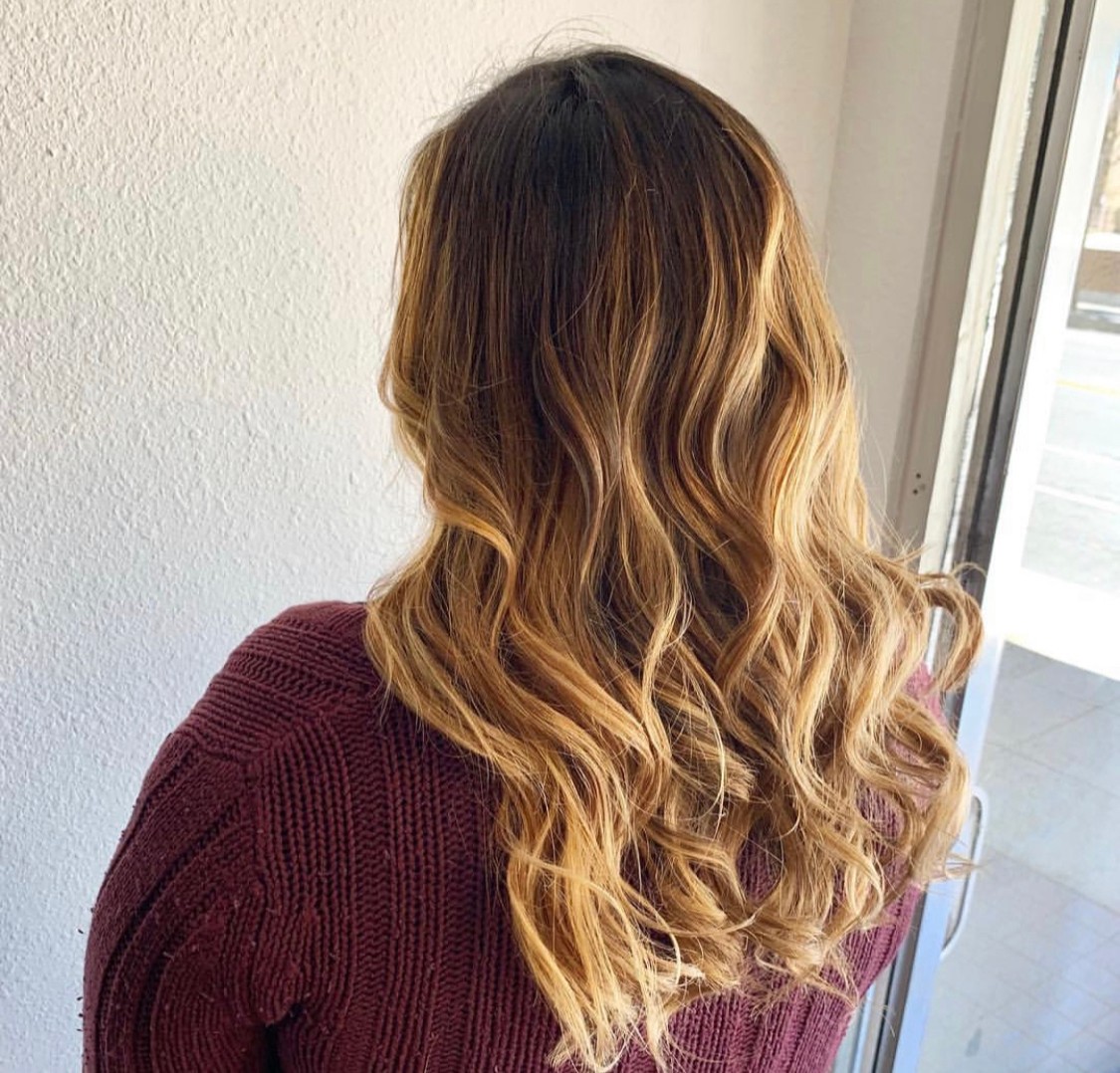 Back view of a woman with long, wavy, ombre hair in shades of brown and blonde, wearing a maroon sweater, near a window.