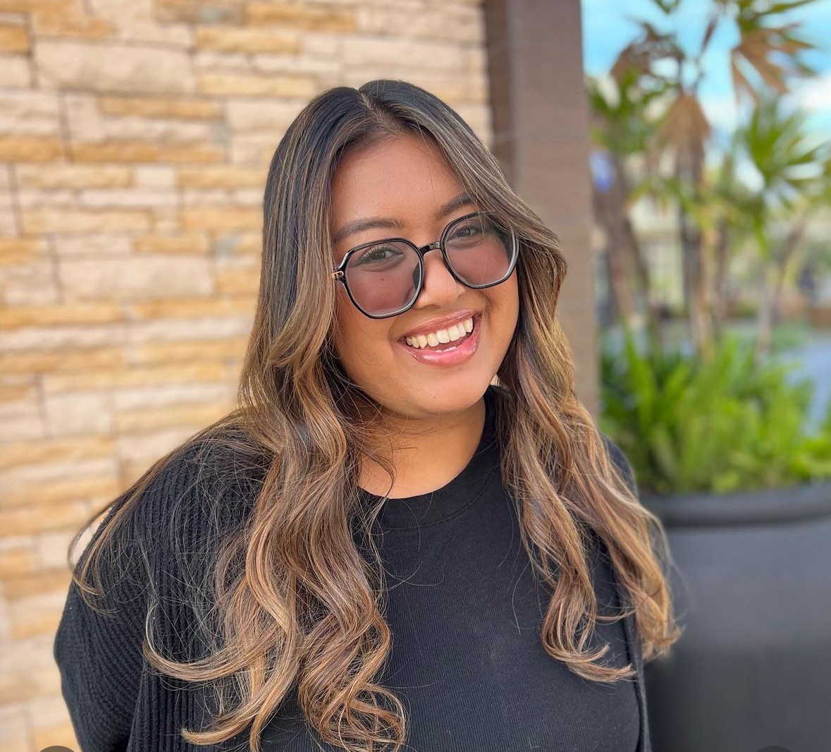 Young woman with long wavy hair, glasses, smiling, wearing a black top, outdoors with plants and a brick wall.