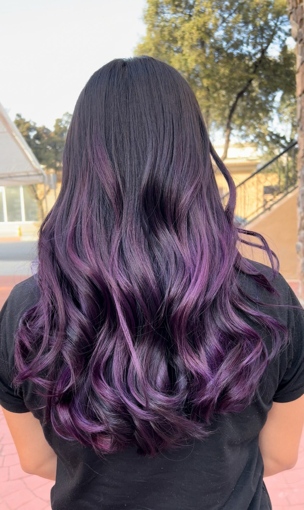 Back view of a person with long, wavy hair dyed purple at the ends, outdoors with trees and a building in background.