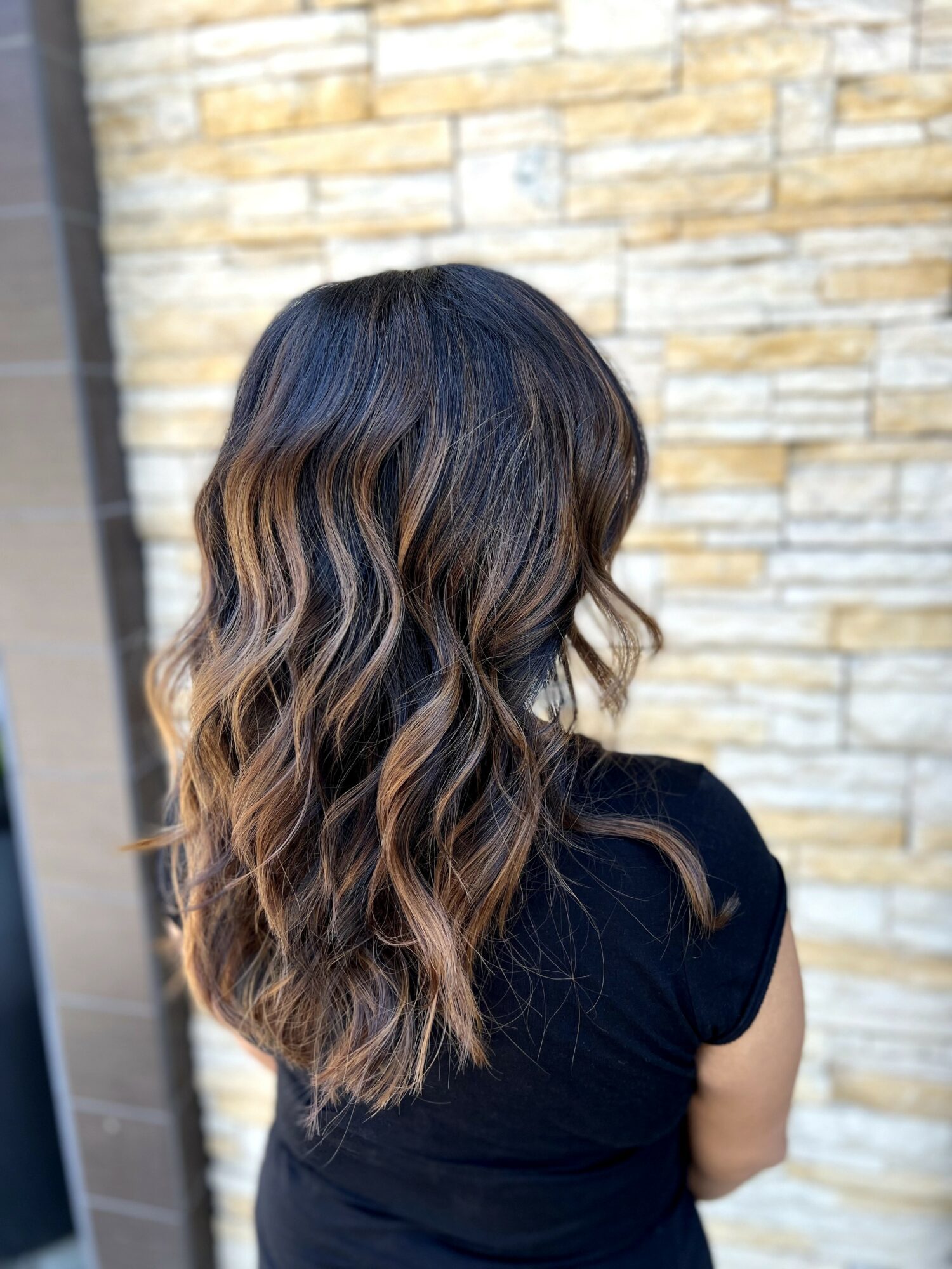 Woman with wavy, ombre hair standing outdoors against a stone wall, facing away from the camera.