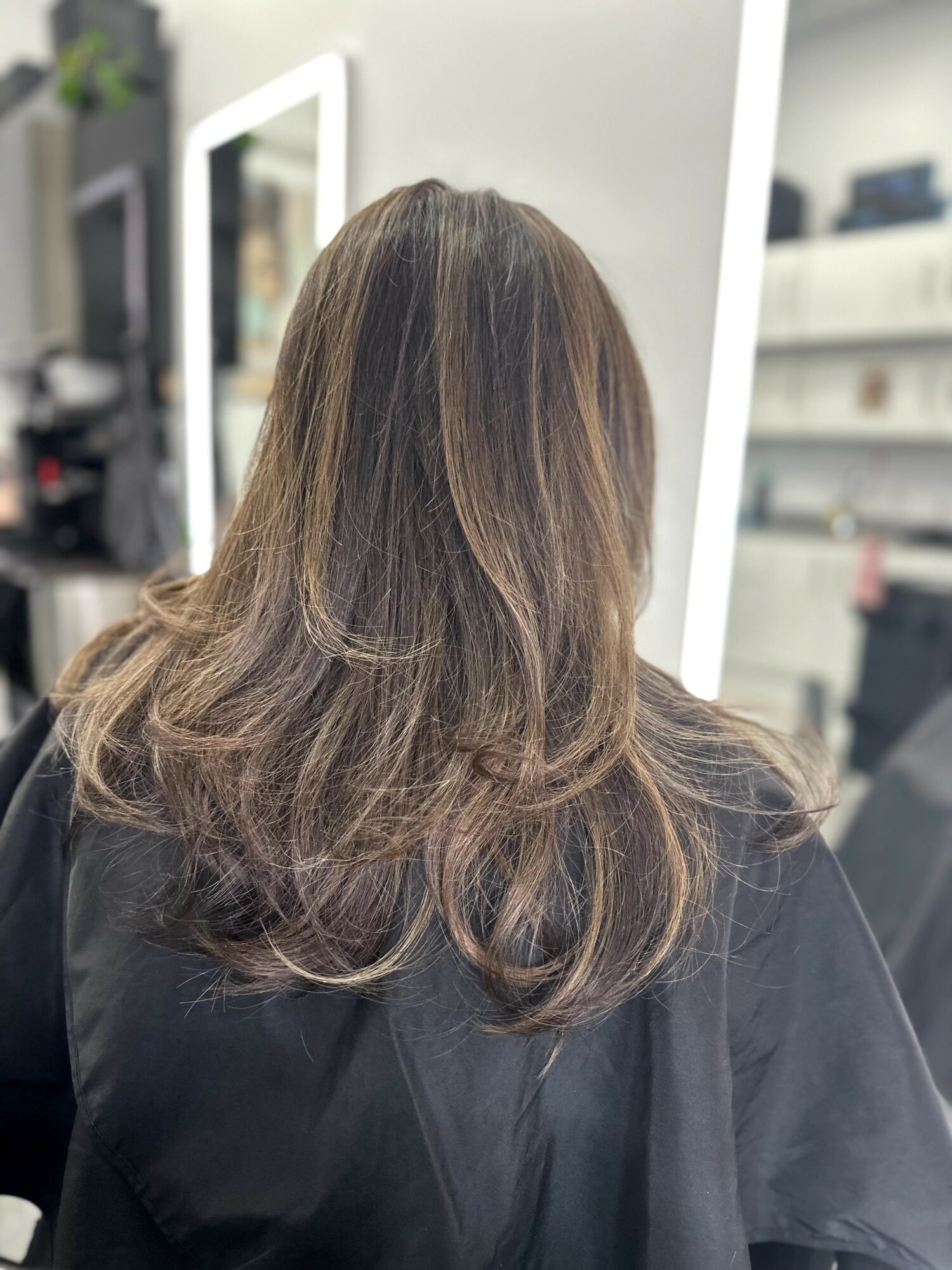 Back of a woman's head with shoulder-length brown hair in a salon, mirrors and shelves in background.
