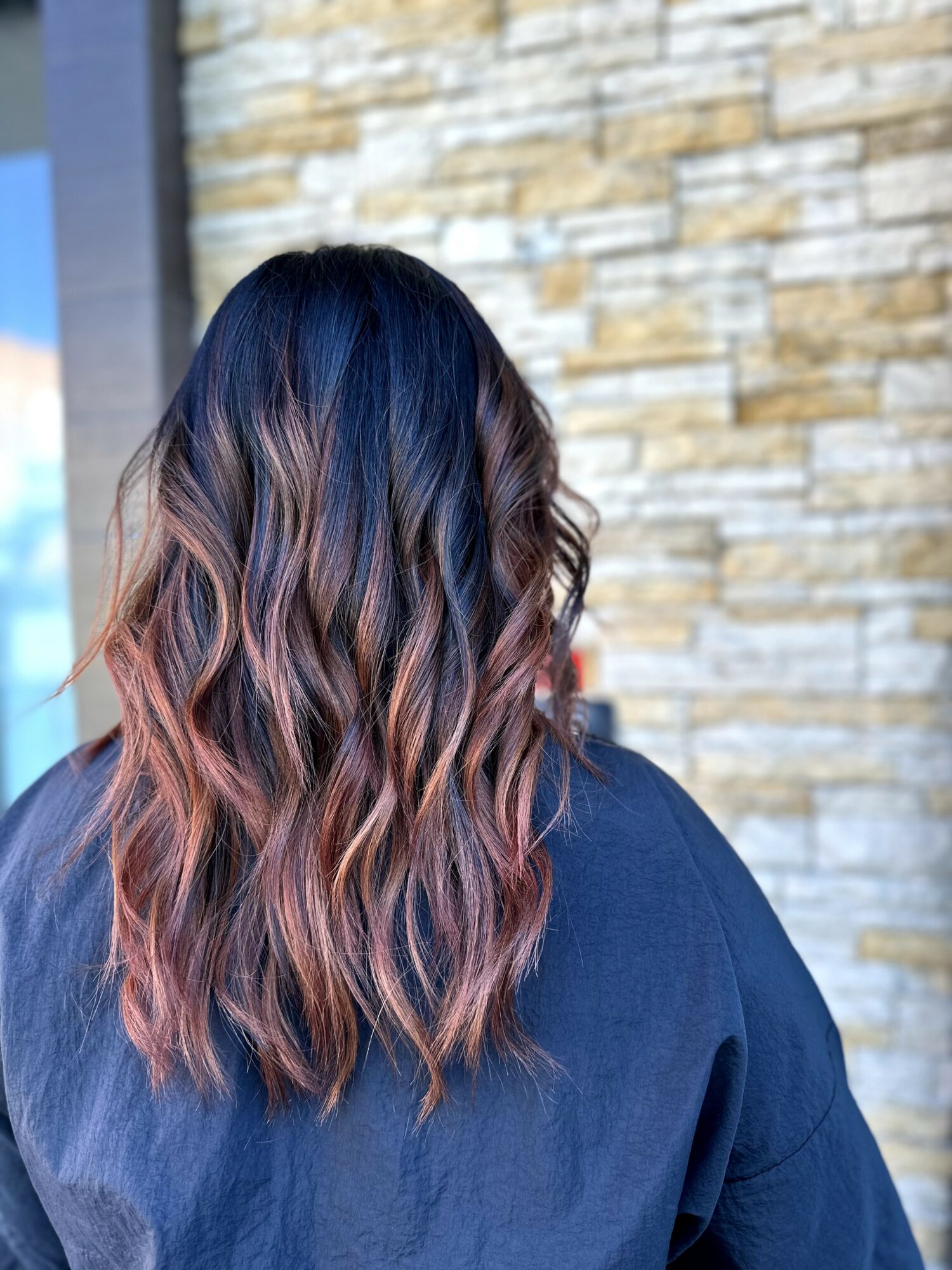 Back of a person with wavy, shoulder-length hair with pink highlights, wearing a dark blue top, standing outdoors near a stone wall.