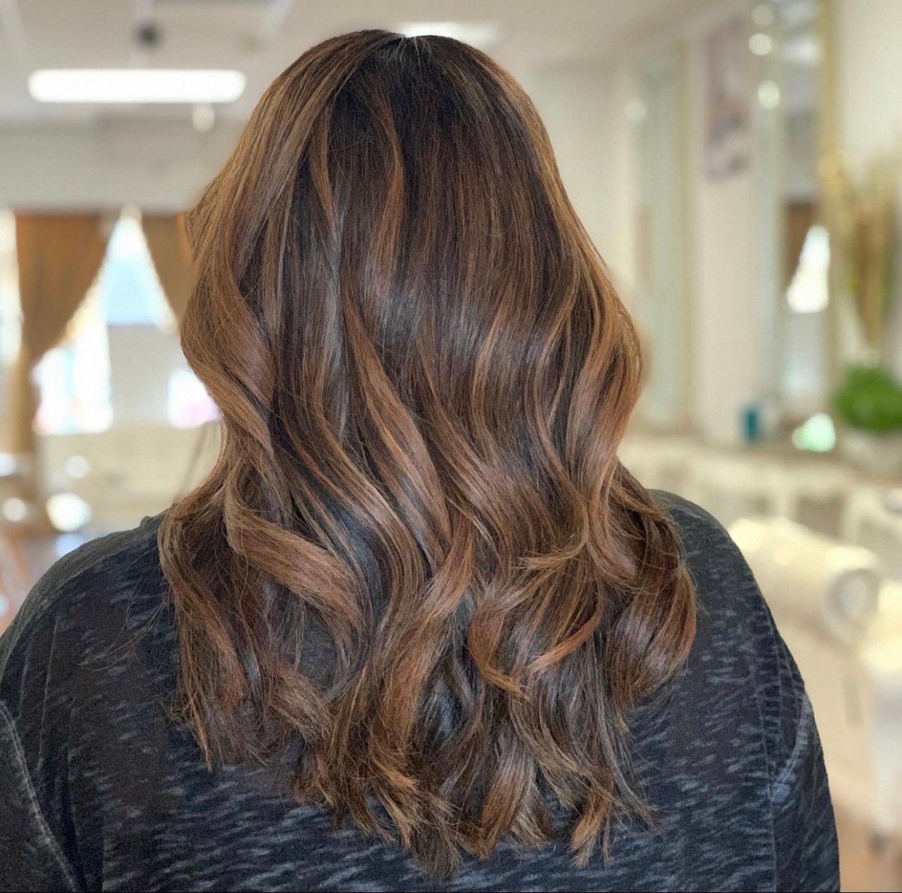 Back view of a person with long, wavy brown hair in a bright room.