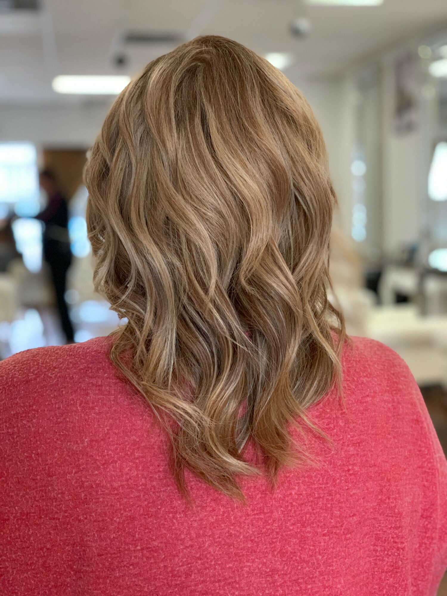 Back view of a woman with wavy, shoulder-length blonde hair wearing a red top in an indoor setting.