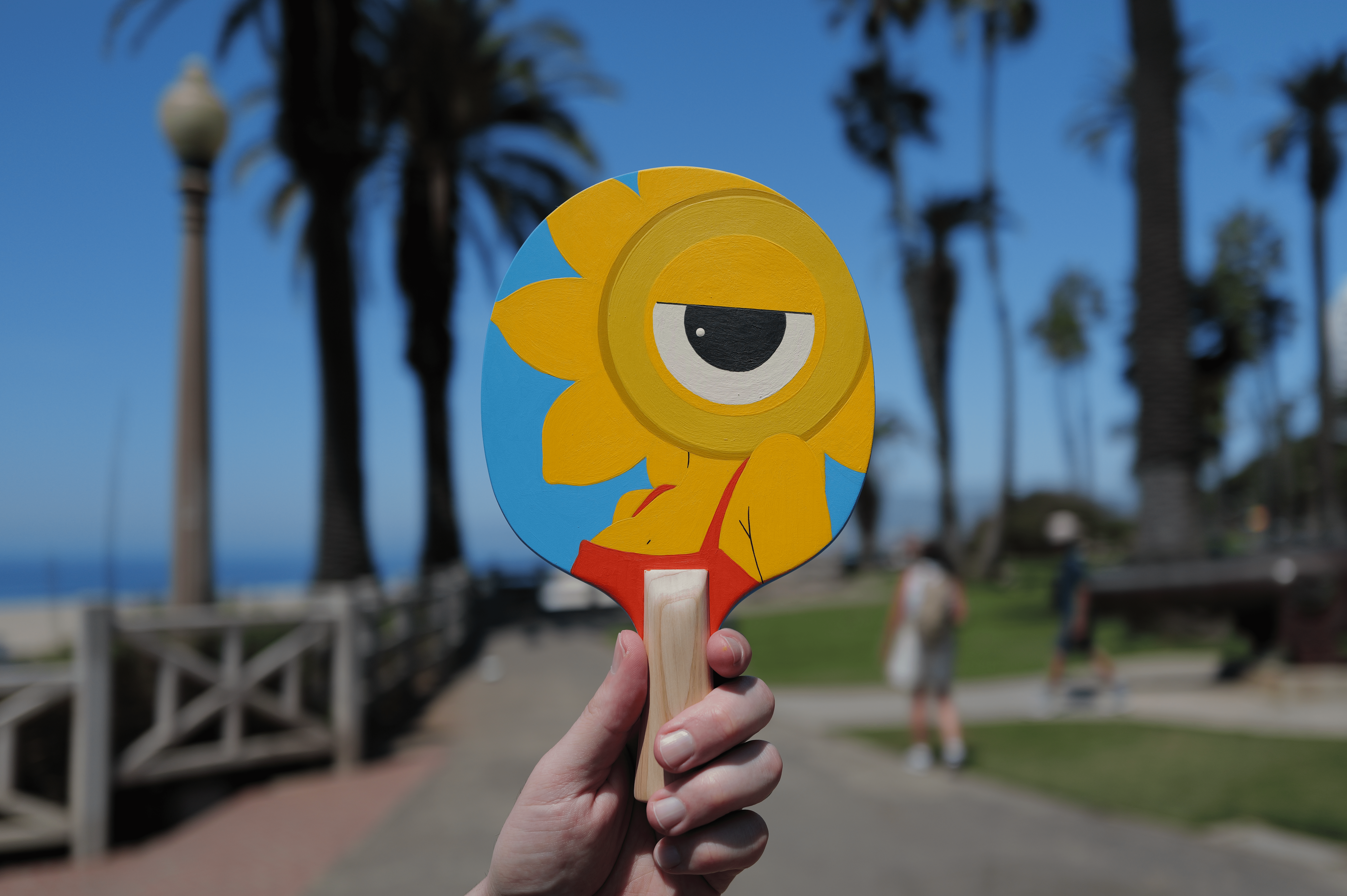 Hand holding a colorful cartoon fish cutout with a background of palm trees and a park, sunny day.