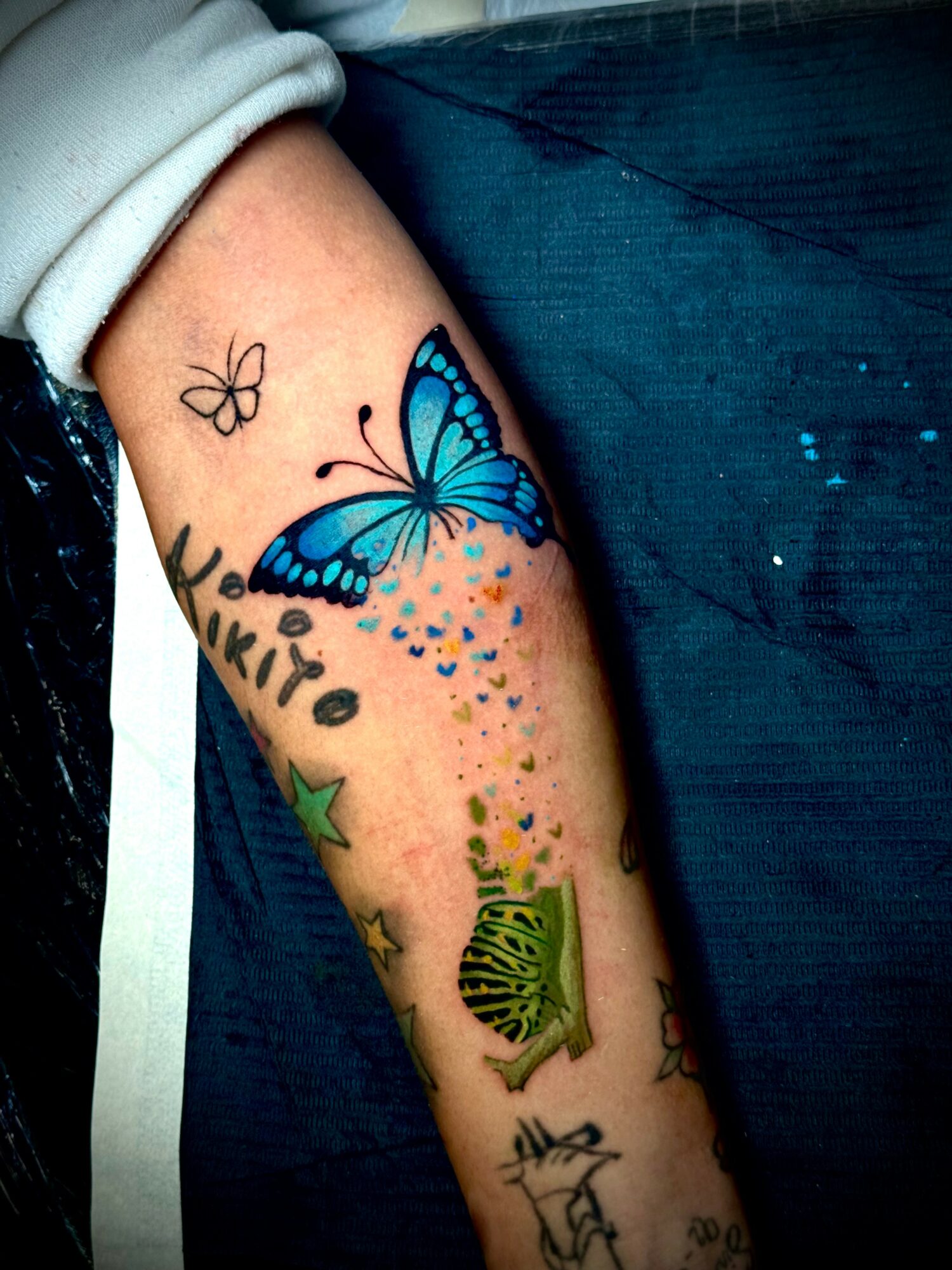 Forearm tattoo featuring a large blue butterfly, smaller butterflies, stars, and decorative elements.