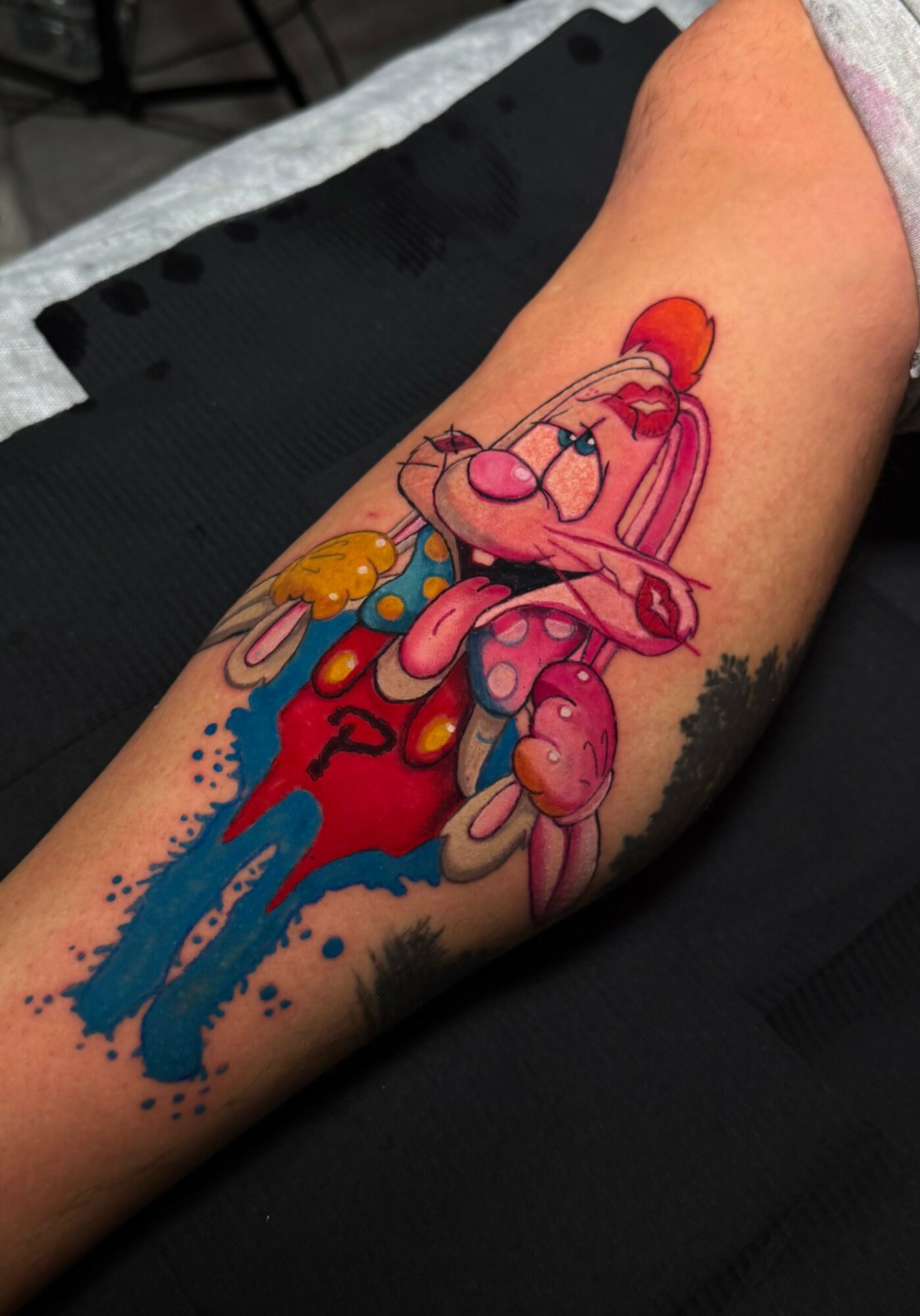 Colorful tattoo of a cartoon rabbit with a red bow, sitting on a blue and red abstract background, on a person's arm.