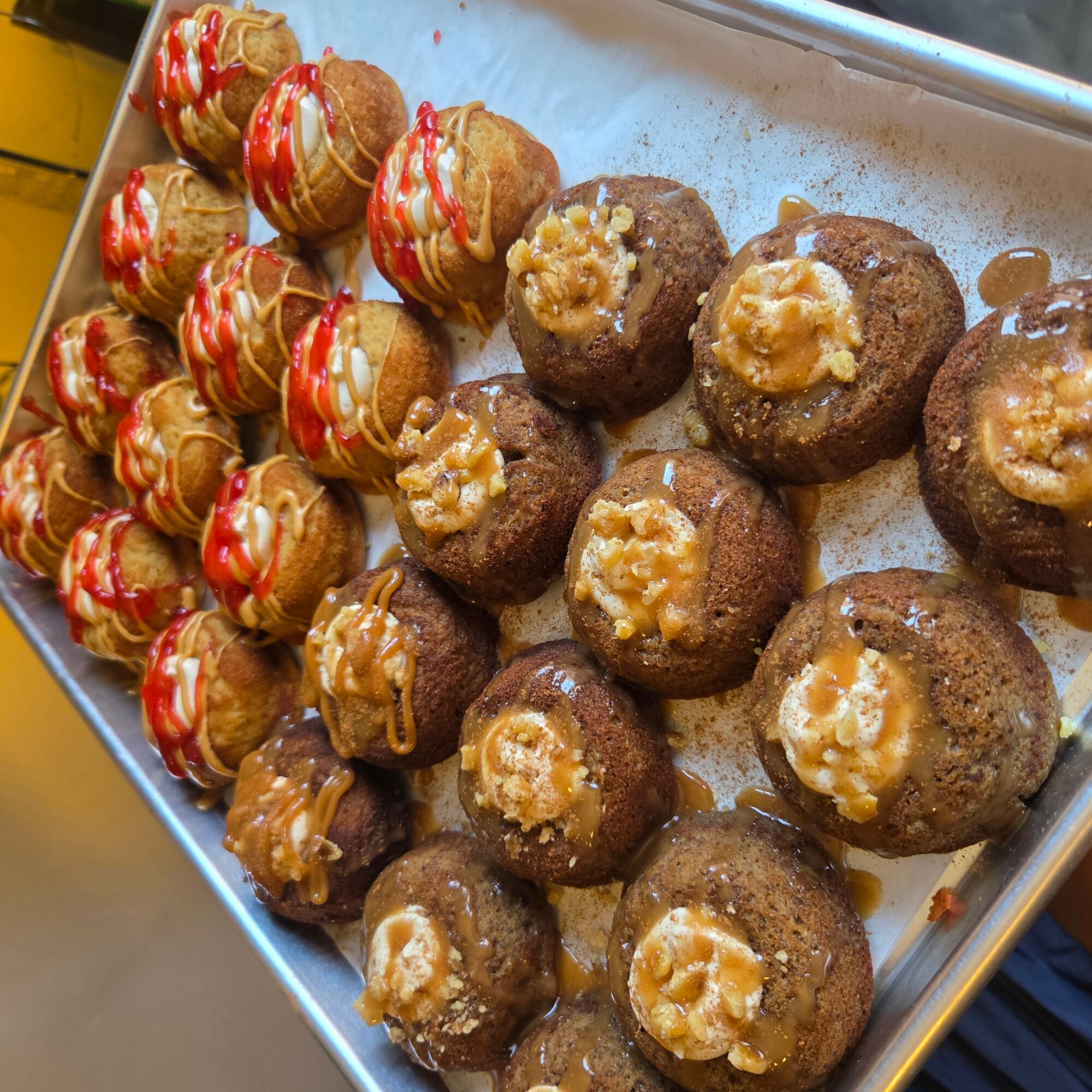 Tray of round desserts topped with caramel and drizzled with red sauce, arranged in rows on a metal tray.