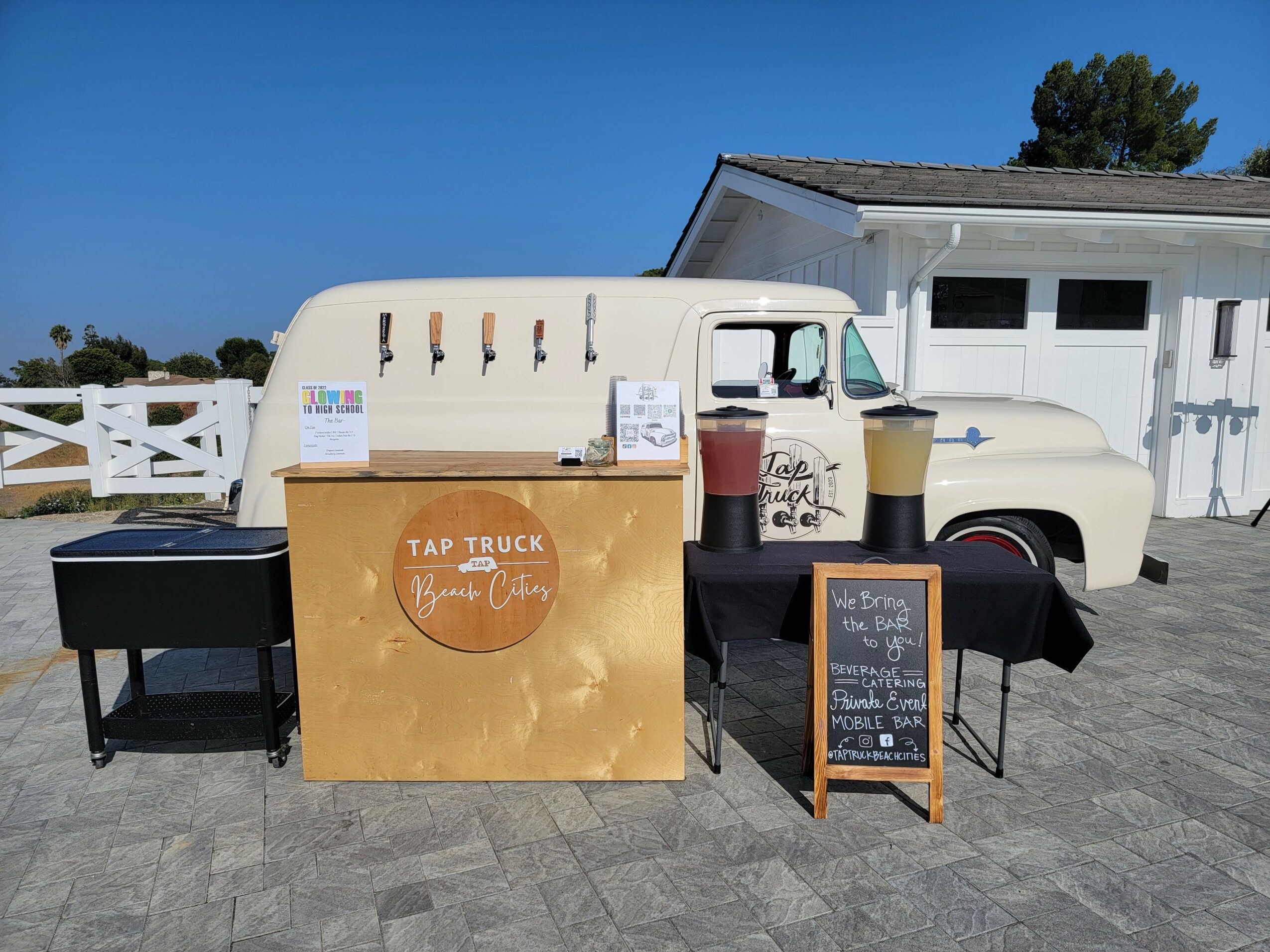 Ice cream truck with a gold sign, black tables, and a chalkboard menu outside a white building under a blue sky.