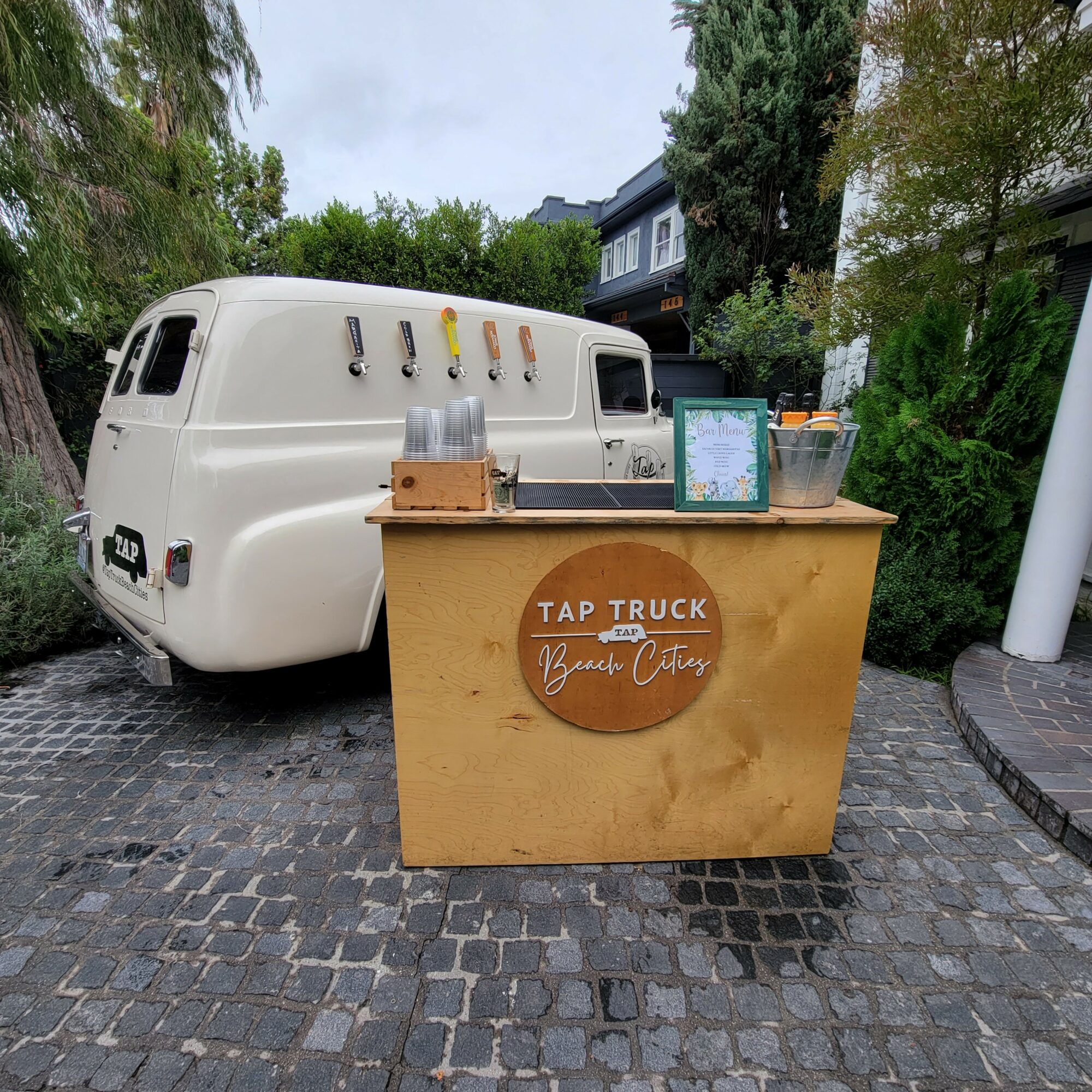 A small food stand with a sign reading 'TAP TRUCK' and a logo, set on a cobblestone street with trees and buildings in the background.