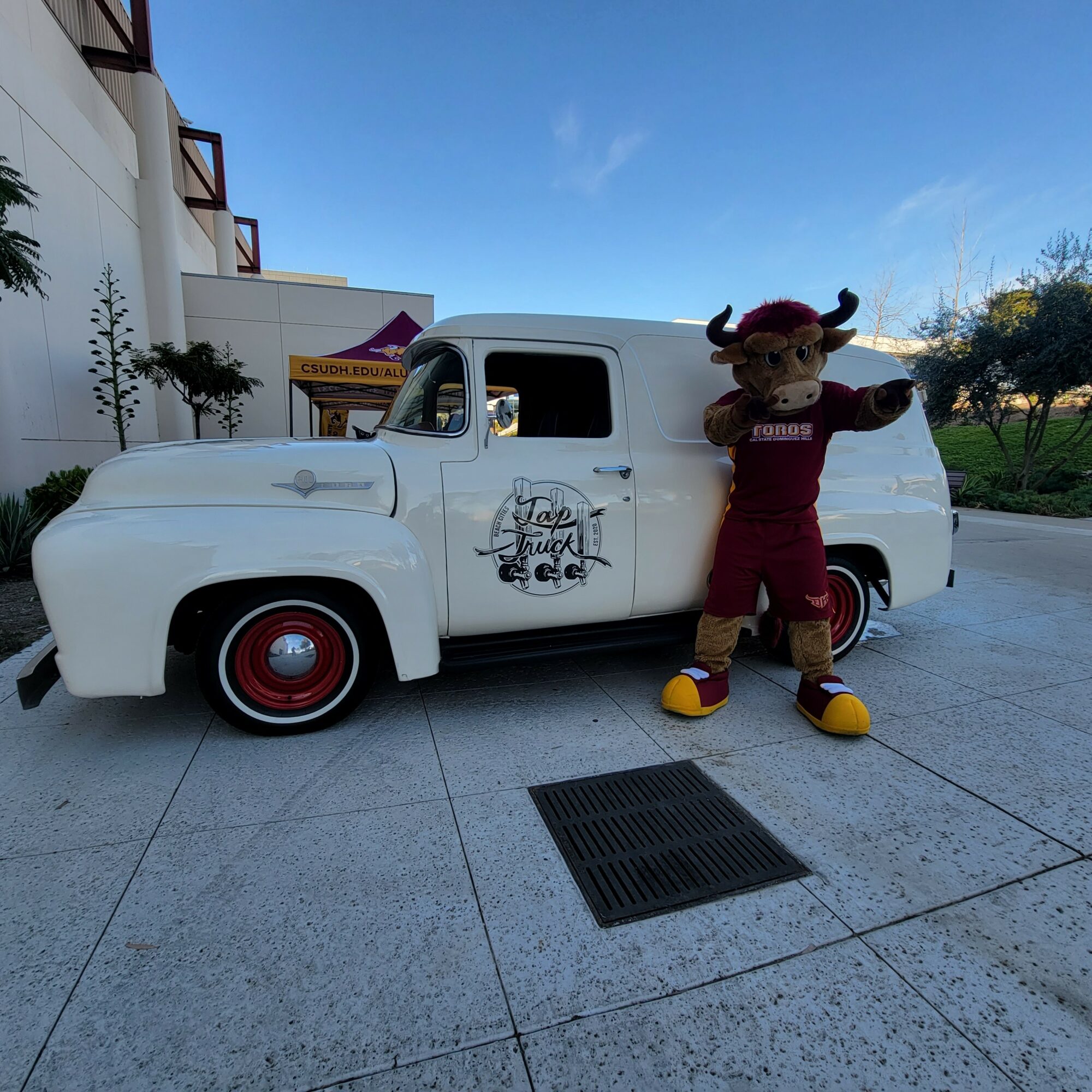 A person in a mascot costume with horns and a tail stands next to a vintage white vehicle with a logo on the door, outdoors.
