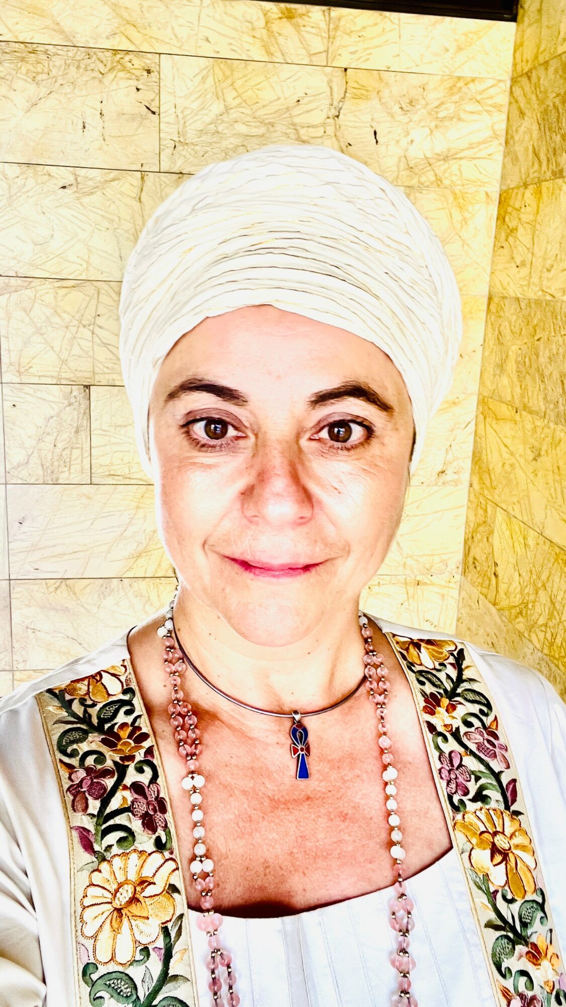 Woman wearing a white headwrap and colorful embroidered clothing, standing against a yellowish stone wall.