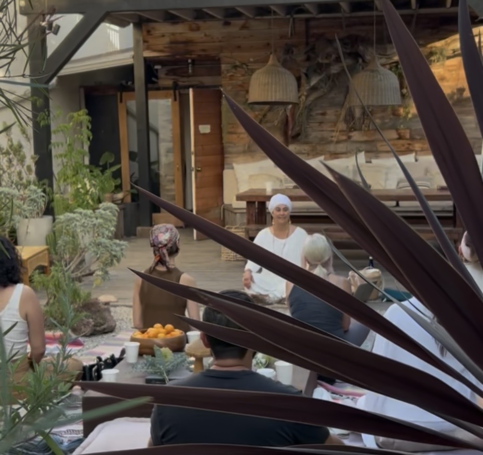 People gathered outdoors, some wearing head coverings, with plants and a wooden structure in the background.
