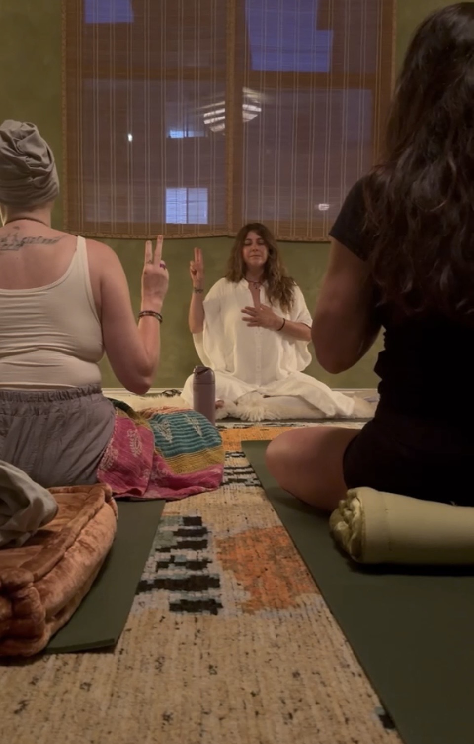 Three people sit on mats in a room, with a woman sitting cross-legged in front of a large window, speaking or meditating.