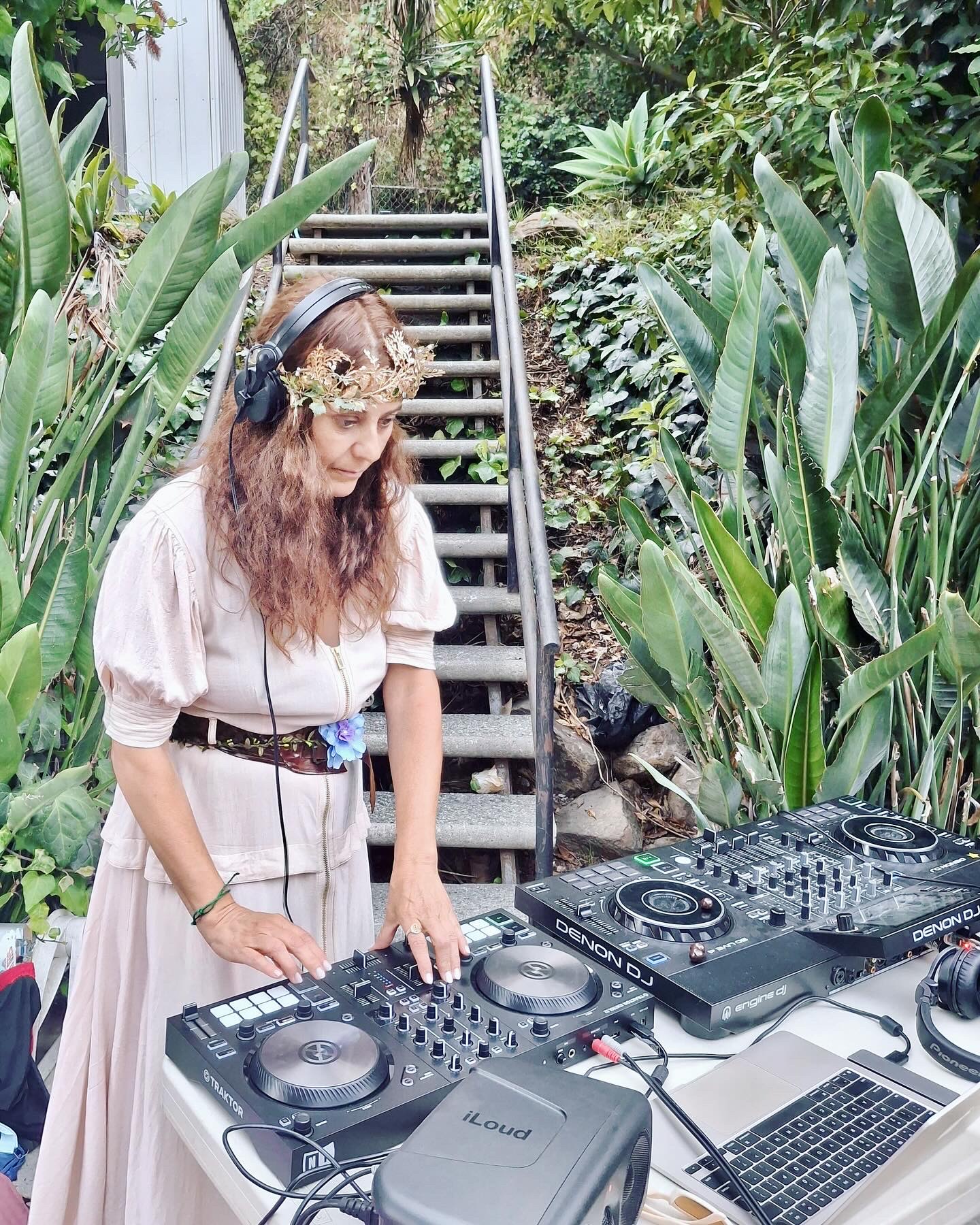 Woman with headphones and floral headband stands near DJ equipment outdoors, surrounded by lush green plants and a staircase.