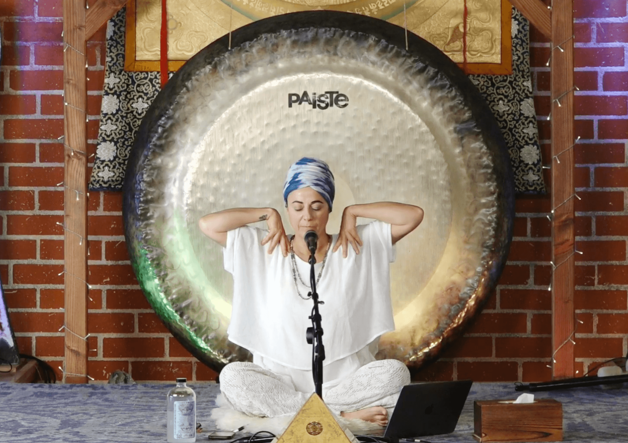 Person sitting cross-legged with hands near ears, in front of a large gong, with brick wall background.