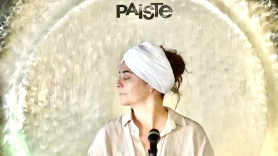 Woman with a white headwrap and beige shirt, standing in front of a textured background, looking to the side.