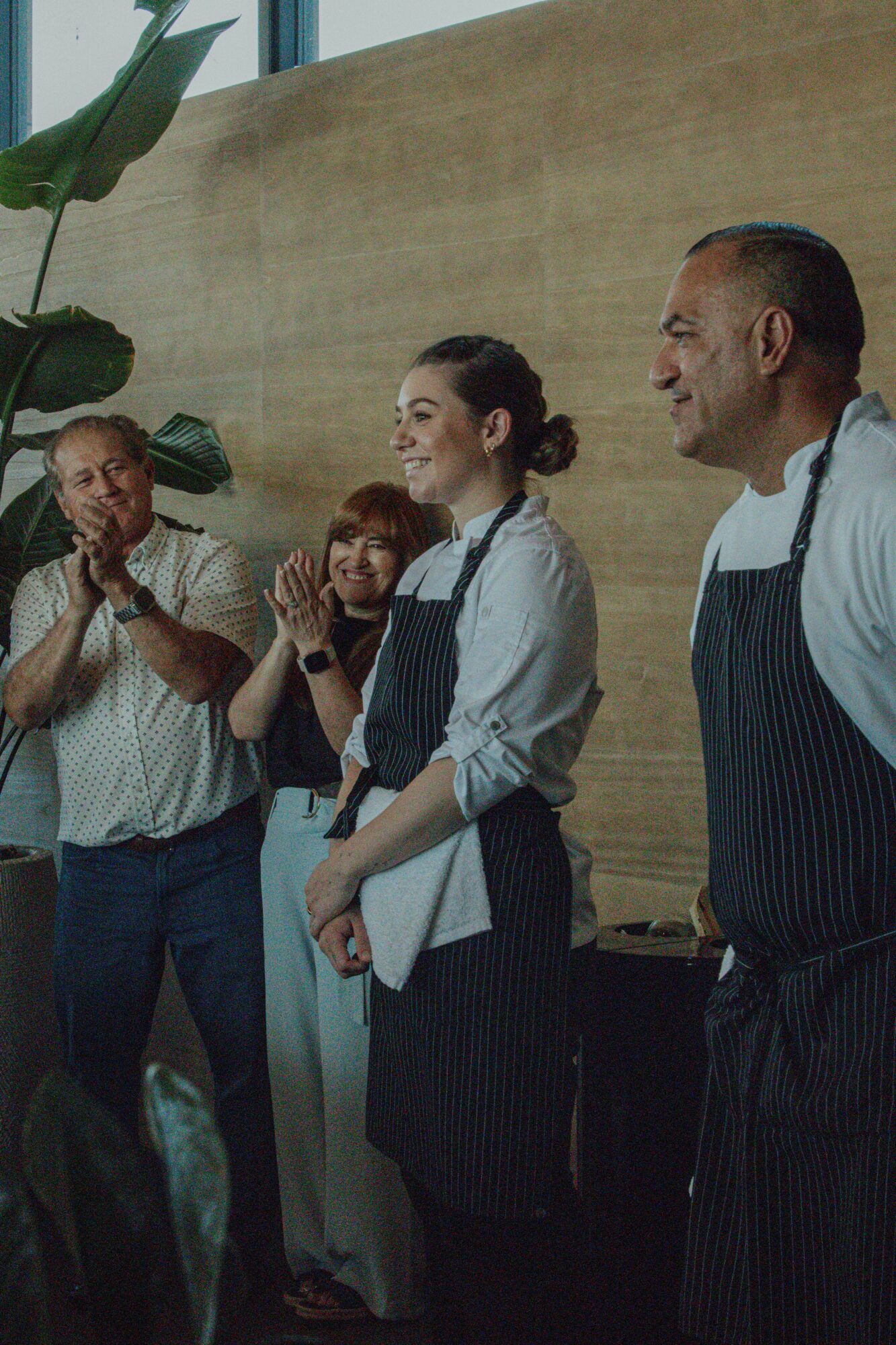 Group of four people in aprons standing indoors, smiling and clapping, with a large plant and plain wall in background.