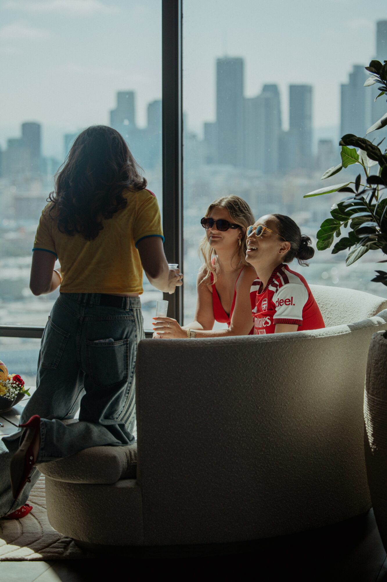 Three women in a room with large window overlooking city skyline, one standing and two sitting, engaged in conversation.