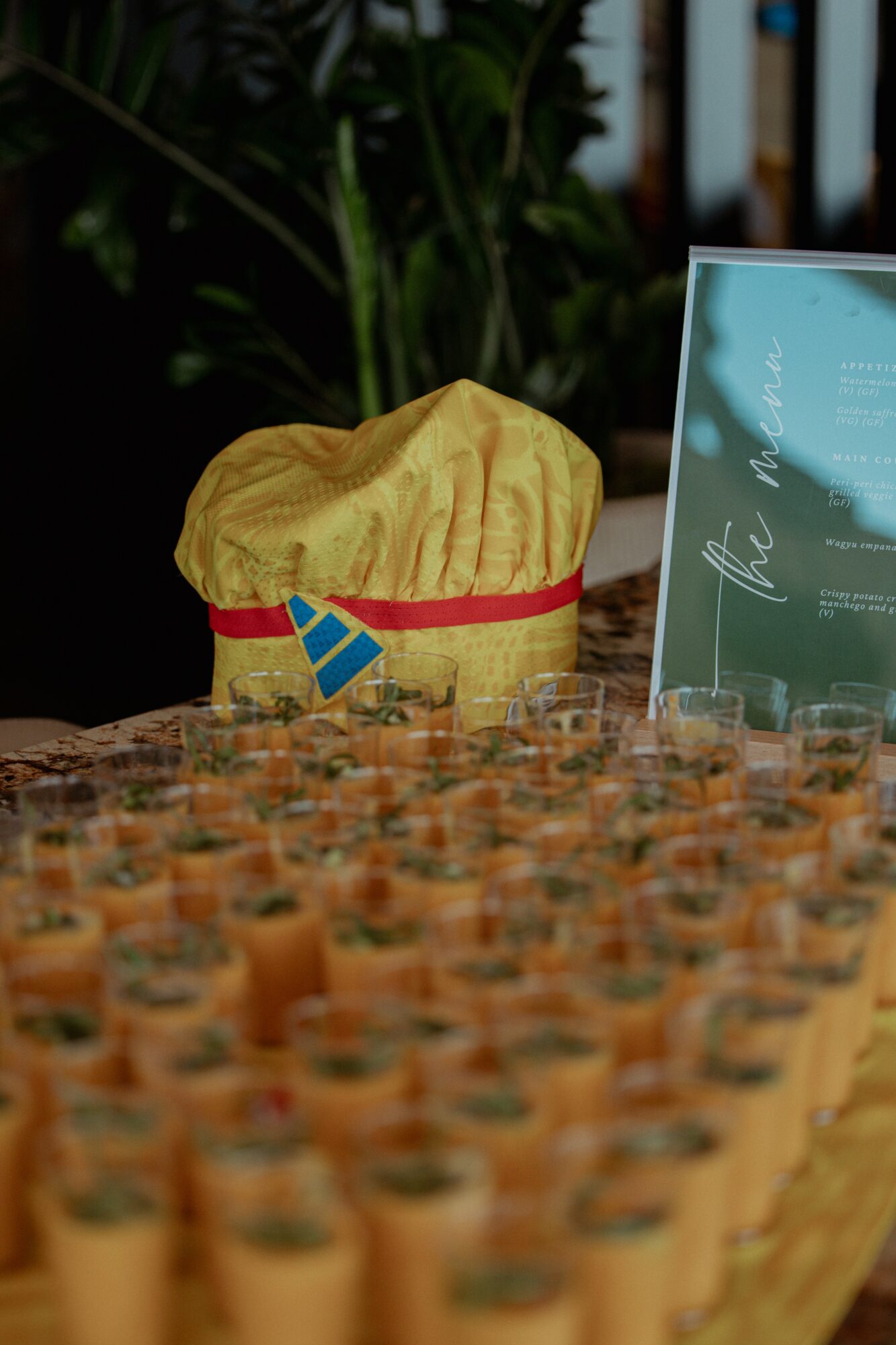 Yellow fabric bag with red trim and a small blue and red flag, placed on a table with many small cups, background with plants and a sign.