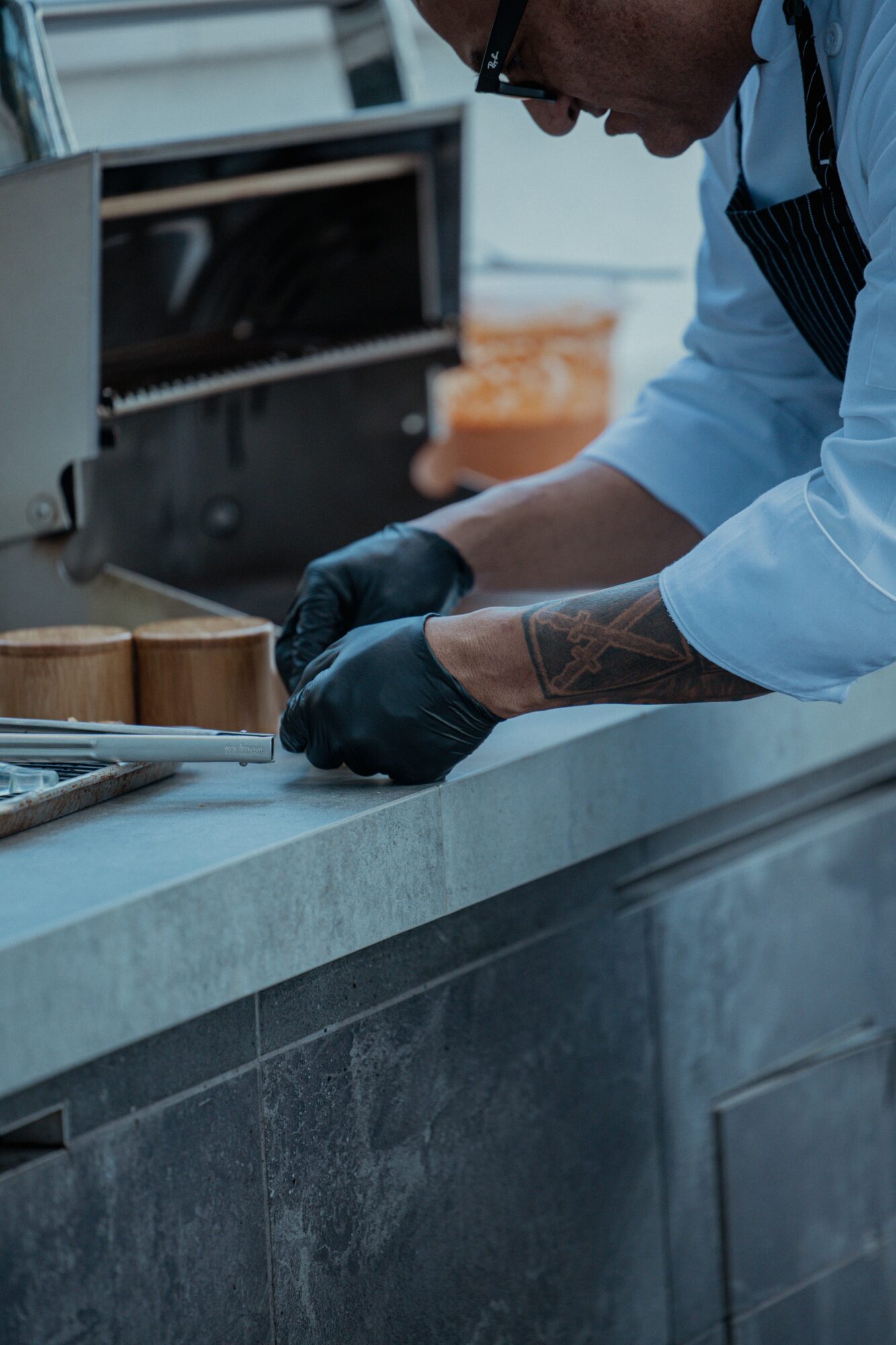 Person wearing black gloves and apron preparing food on a kitchen counter, with a grill in the background.