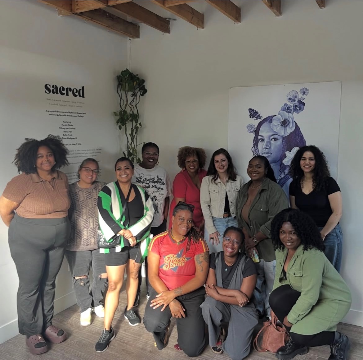 Group of ten diverse women standing and sitting indoors, with artwork and a plant on the wall behind them.