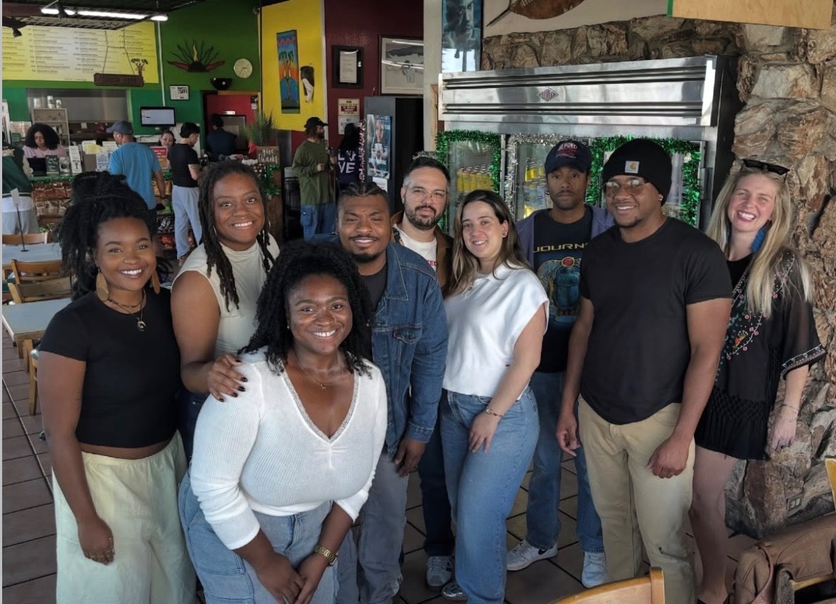 Group of ten people smiling indoors, some standing and some sitting, in a colorful restaurant or cafe setting.