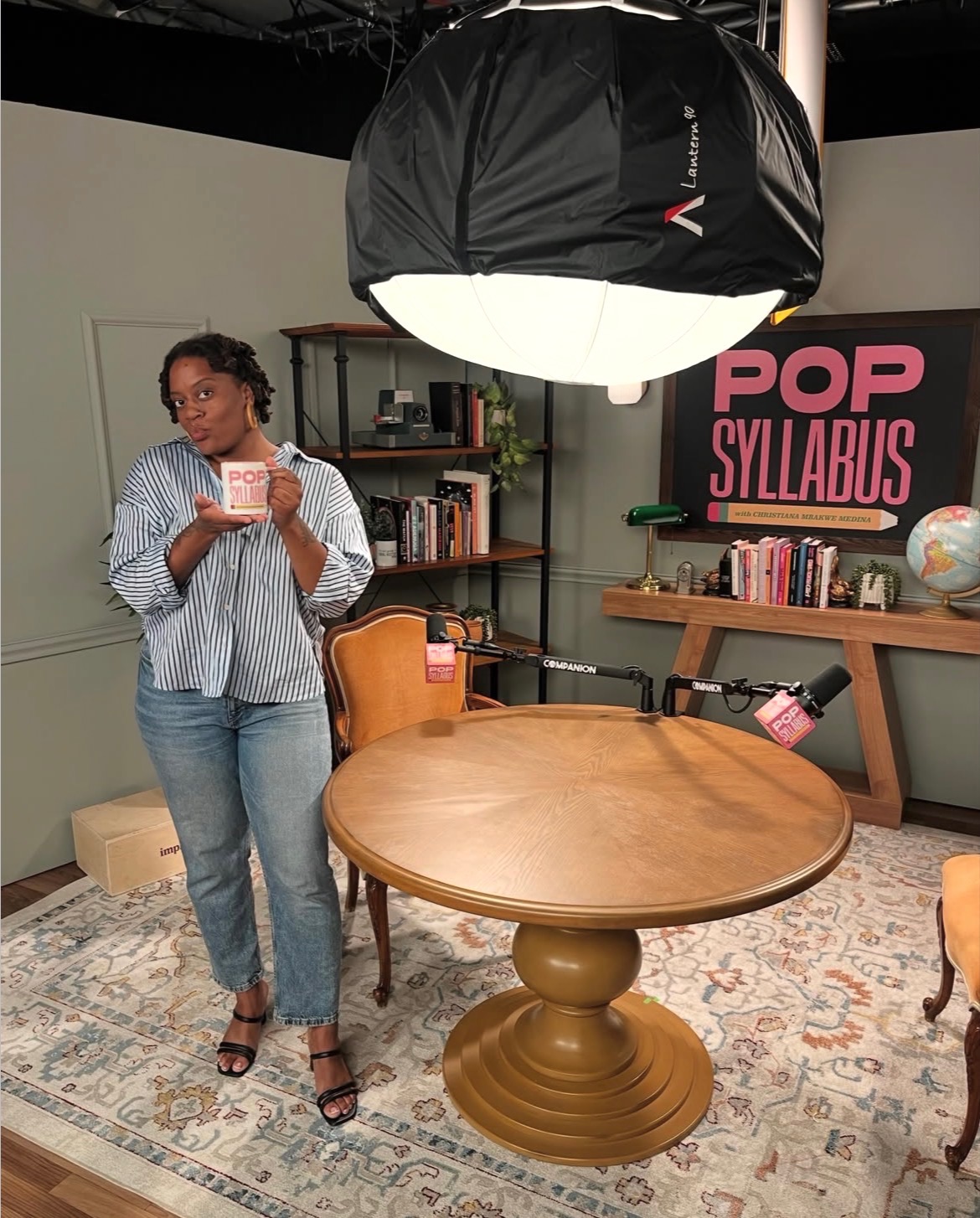 Woman taking a selfie in a room with a round wooden table, bookshelf, and a pink sign that says 'POP SYLLABUS'.