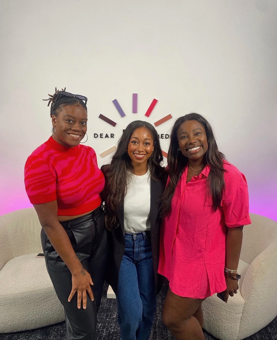 Three women smiling, standing close together in front of a white wall with a logo, two sofas behind them.