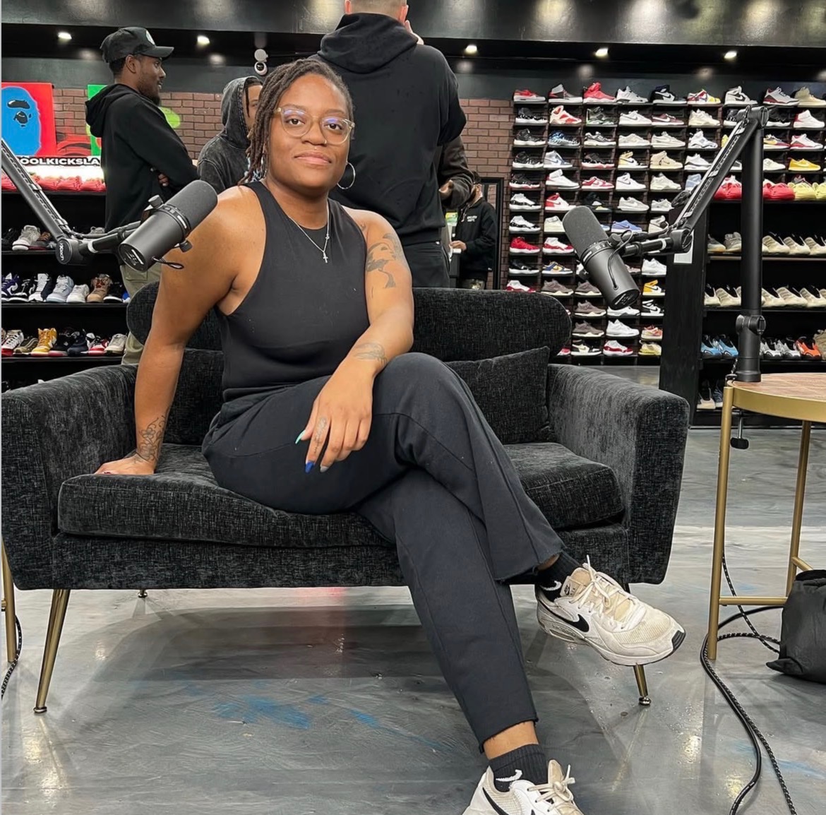 Woman sitting on a black sofa with microphones, in a shoe store, wearing glasses and a black outfit.