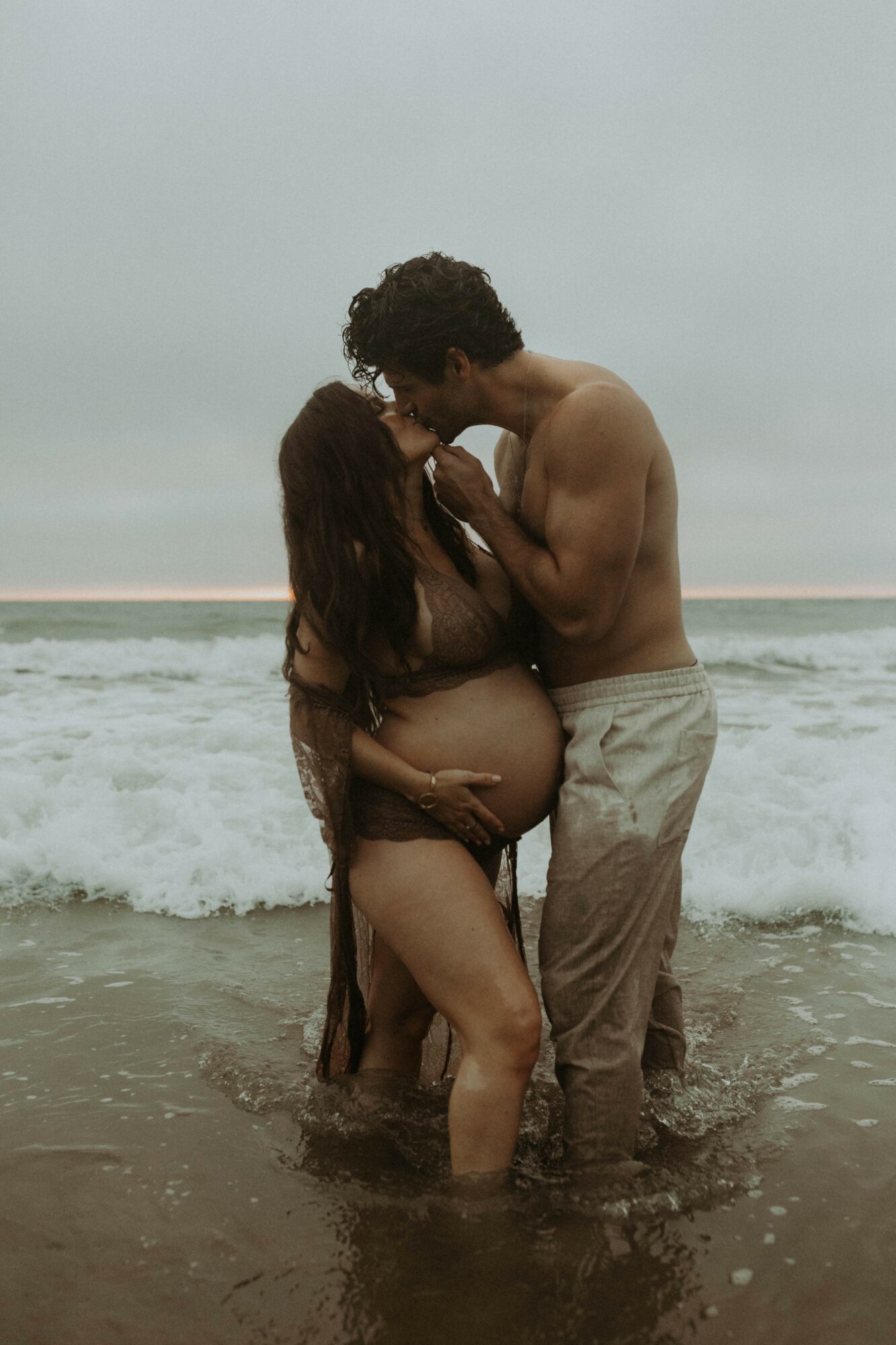 A pregnant woman and a man stand in the ocean, embracing and touching foreheads, with waves in the background.