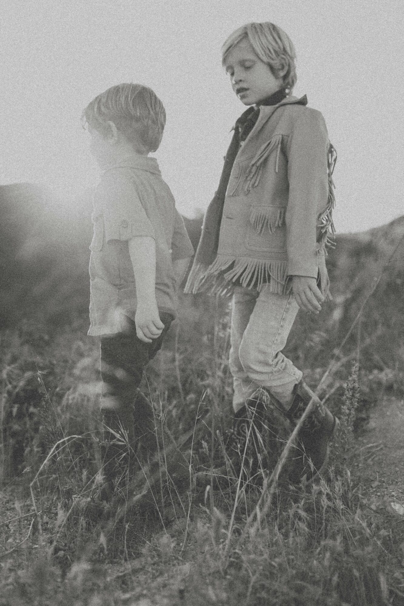 Two children standing in a grassy field, one older and one younger, with a natural outdoor background, in black and white.