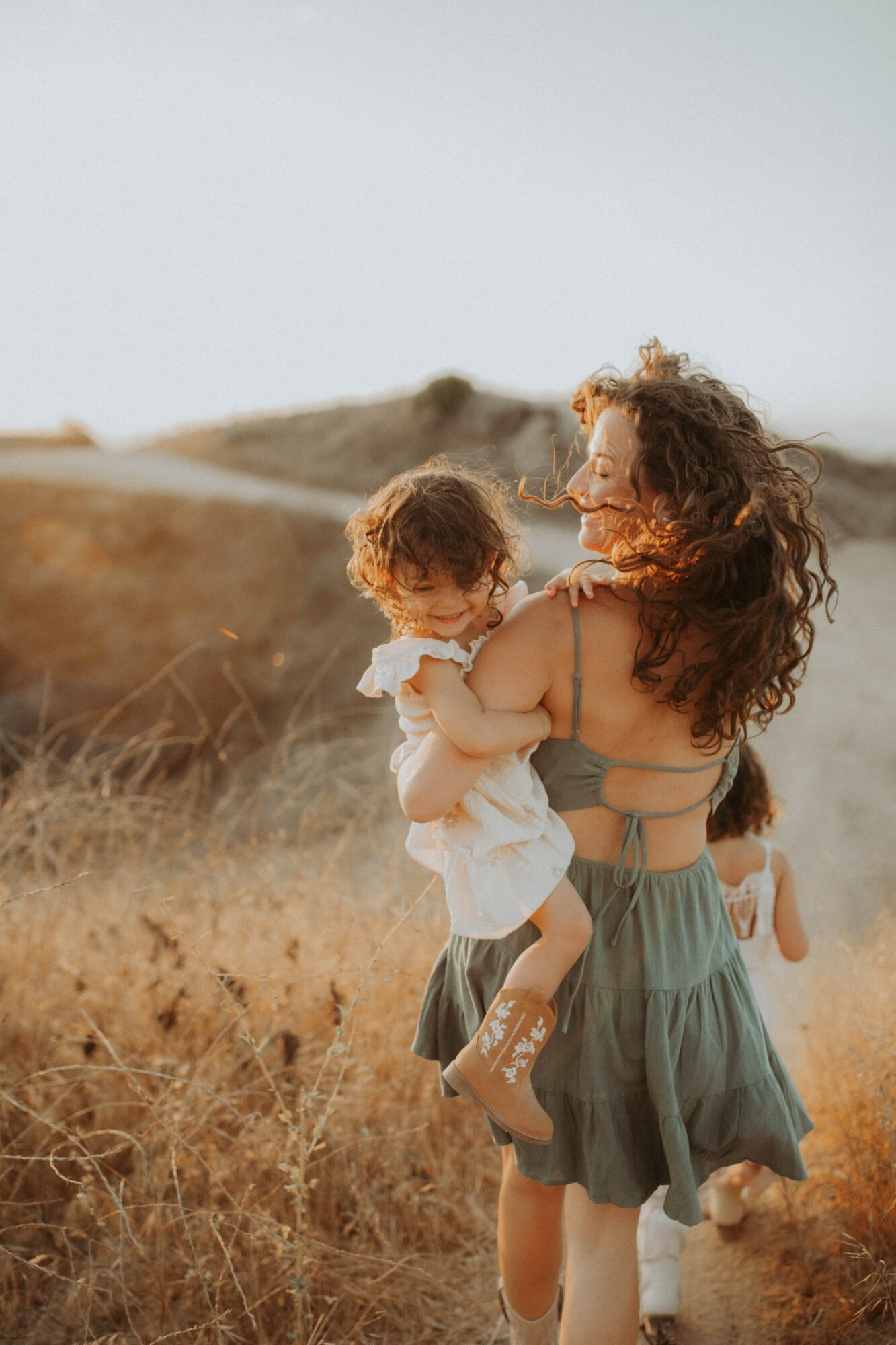 Woman holding a child in a field with dry grass, hills, and a clear sky in the background.