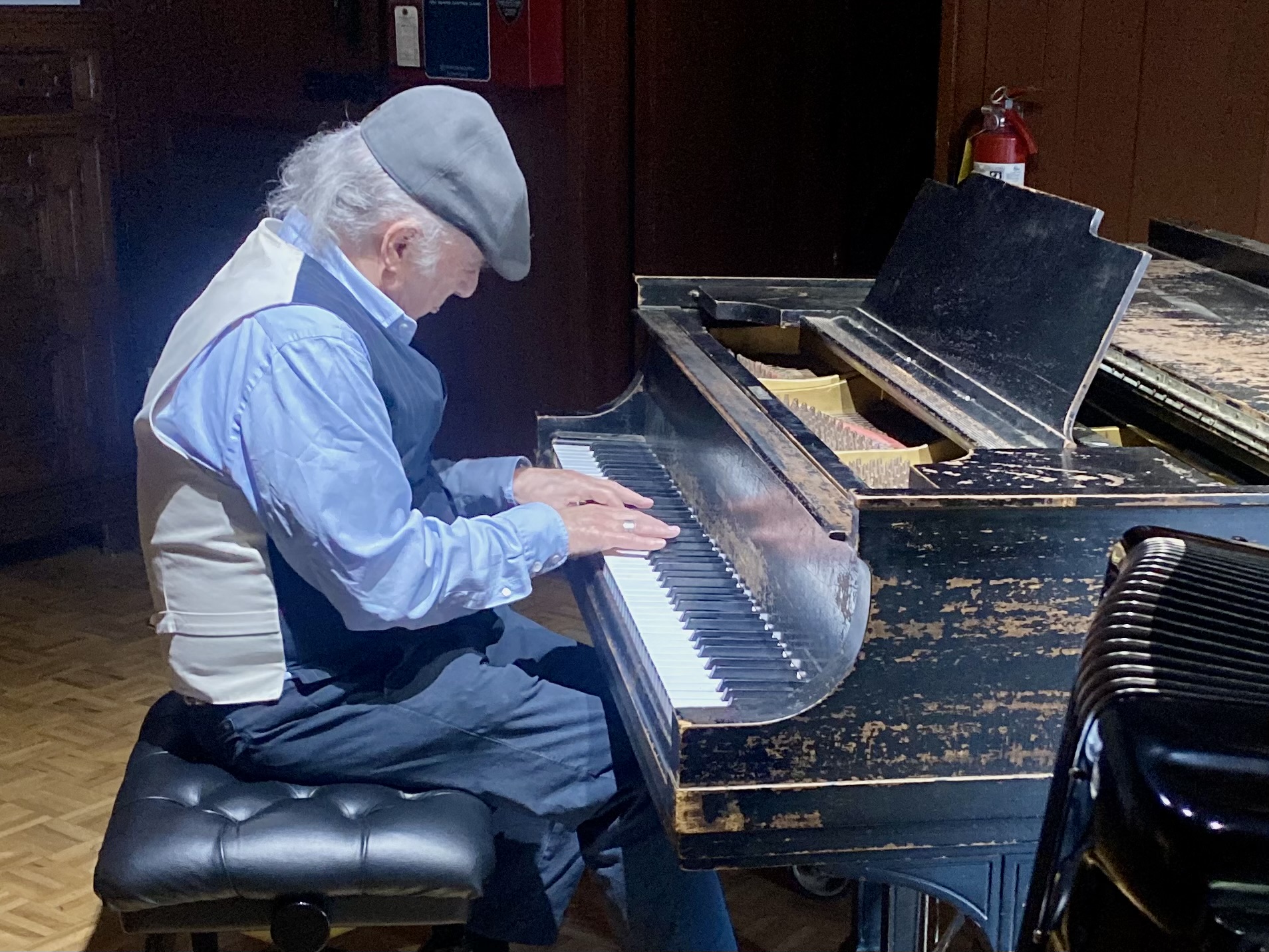 Elderly man playing a worn black grand piano, sitting on a black cushioned bench, in a wooden room.