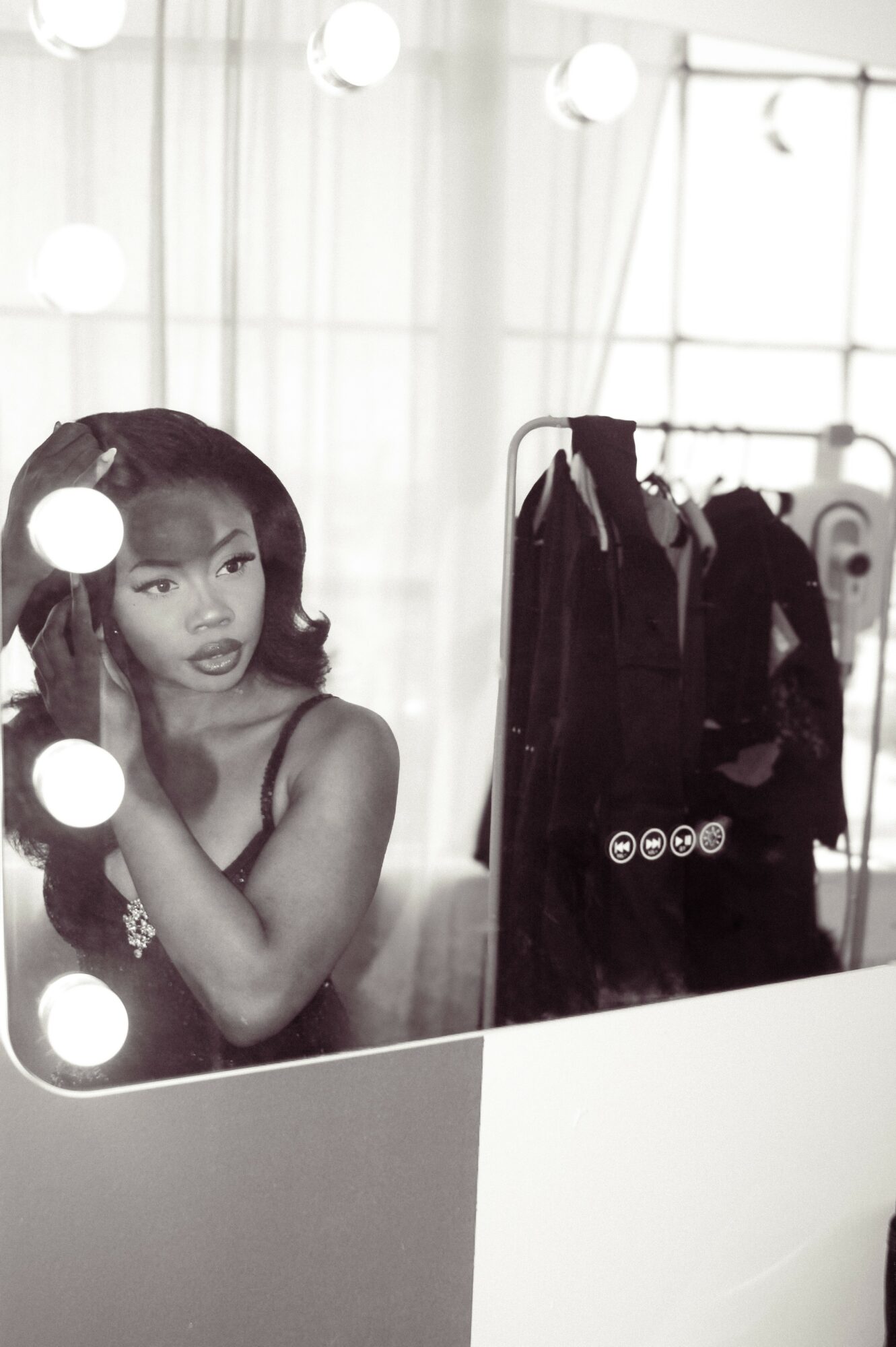 Black and white photo of a woman with wavy hair looking at a mirror, with clothing on a rack in the background.