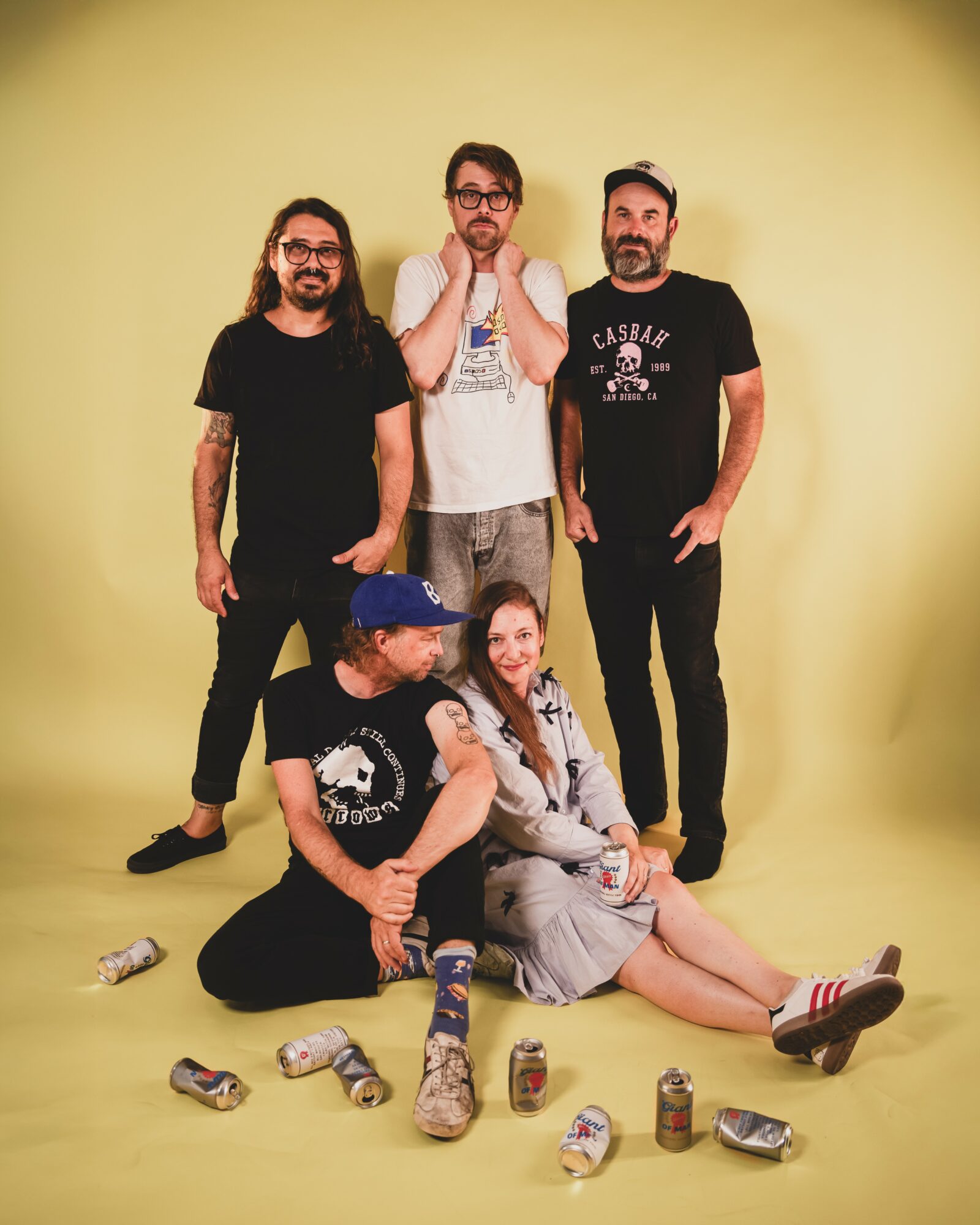 Five people, three standing and two sitting, with cans on the floor, against a yellow background.