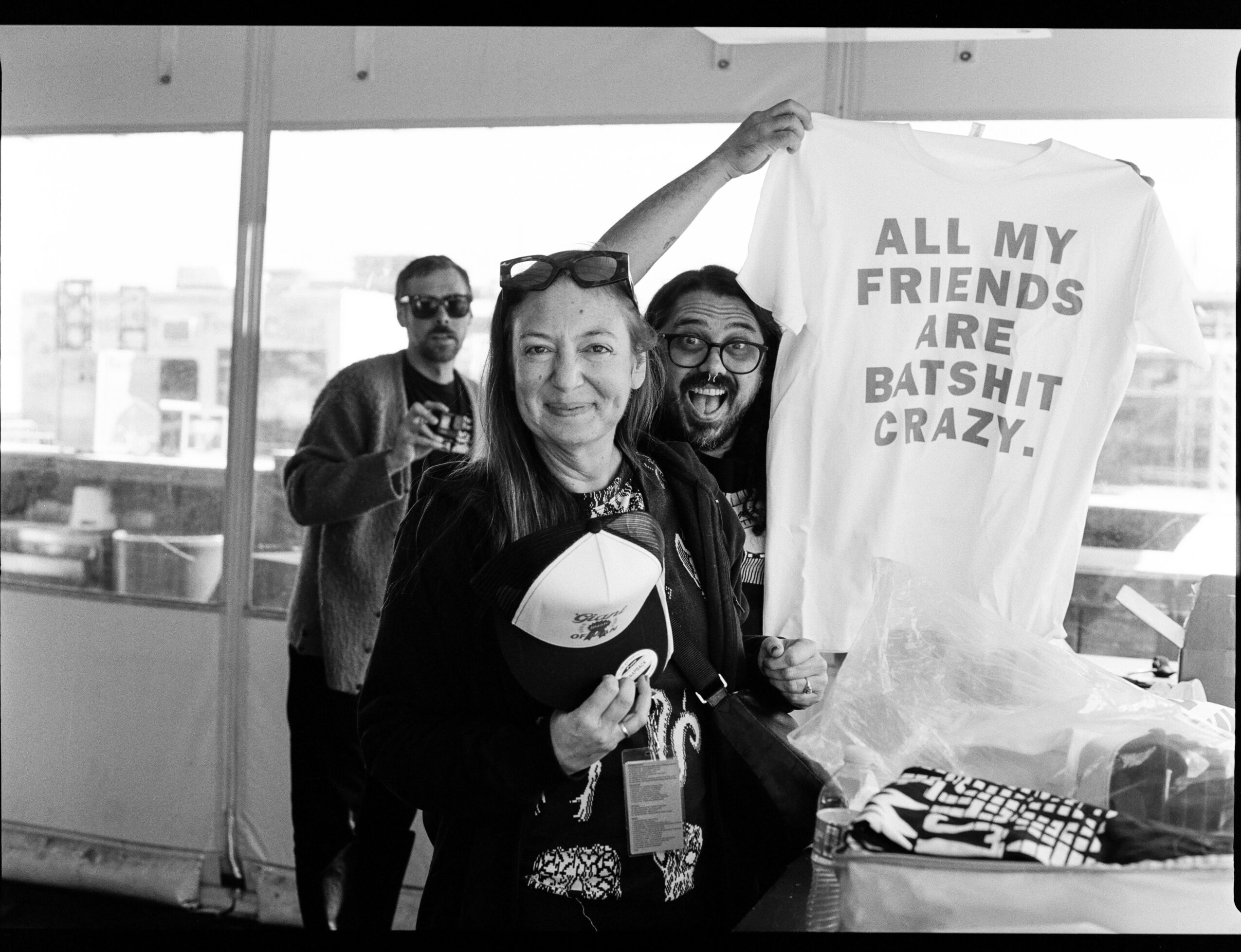 Three people at a gathering, one holding a sign that reads 'All my friends are batshit crazy,' with a city view in the background.