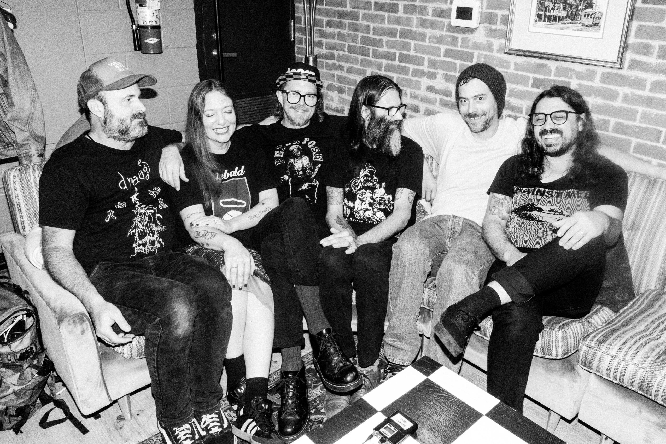 Group of six people sitting on a couch and chairs, smiling and talking, in a room with brick walls.