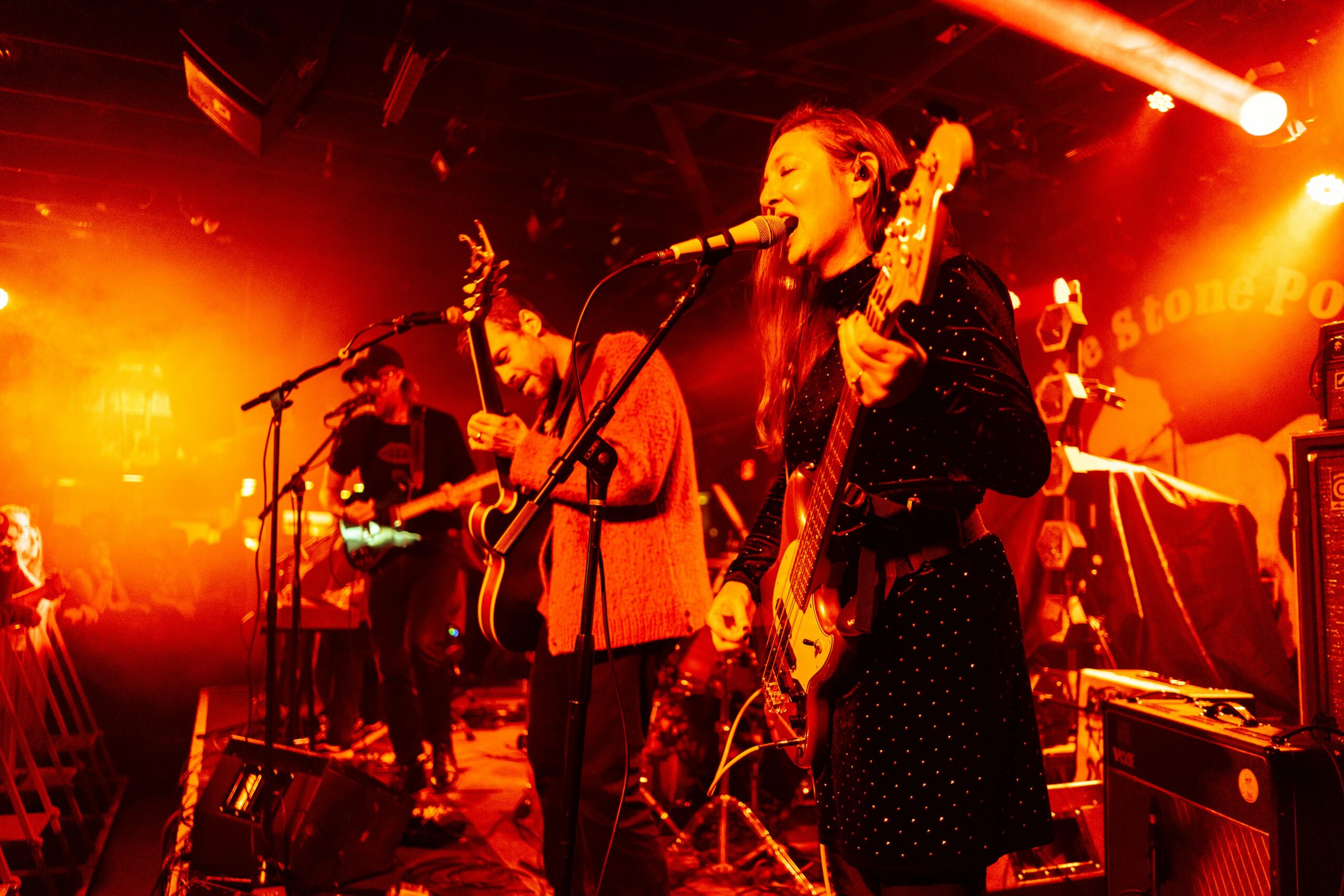 Band performing on stage with bright orange lighting, singer playing guitar and singing into microphone, other musicians in background.