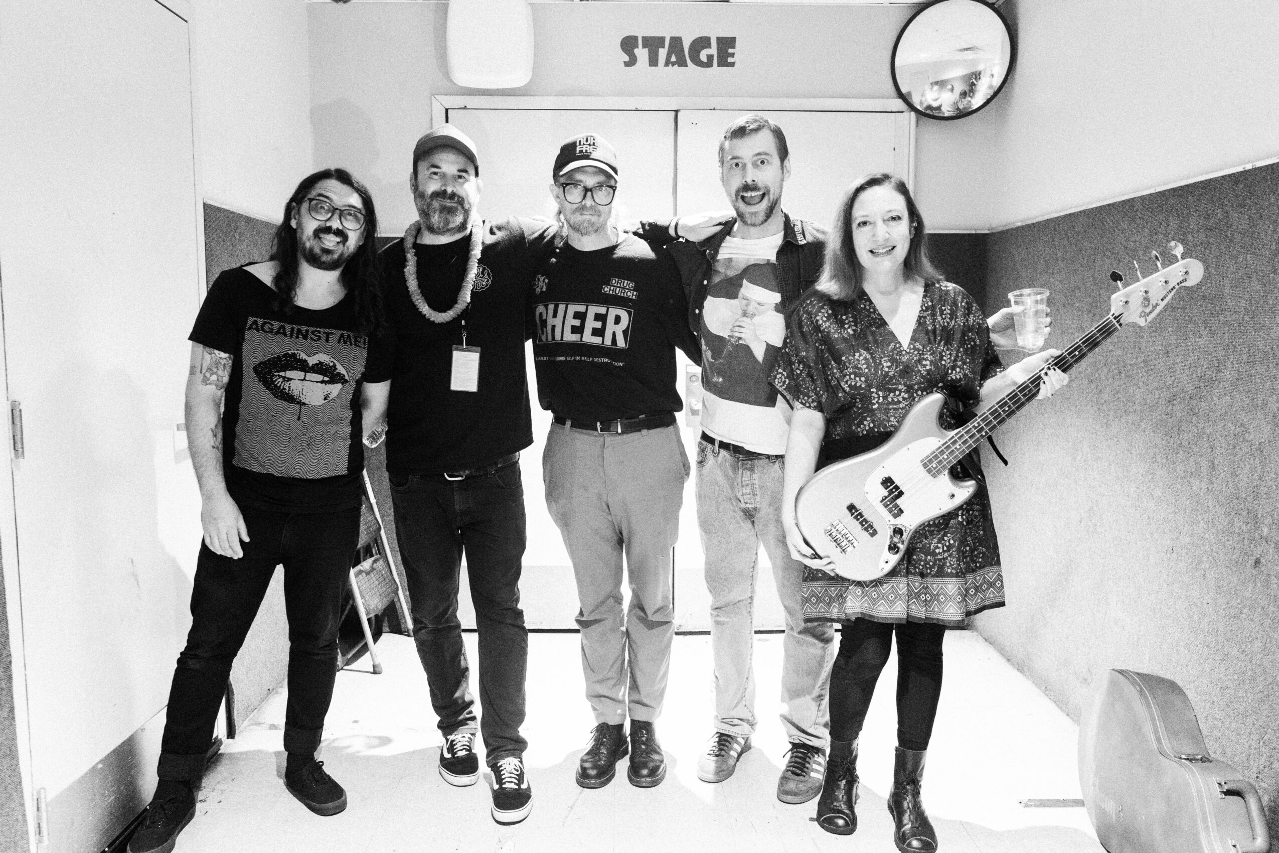 Group of five people standing in front of a stage, smiling, with one person holding a bass guitar.
