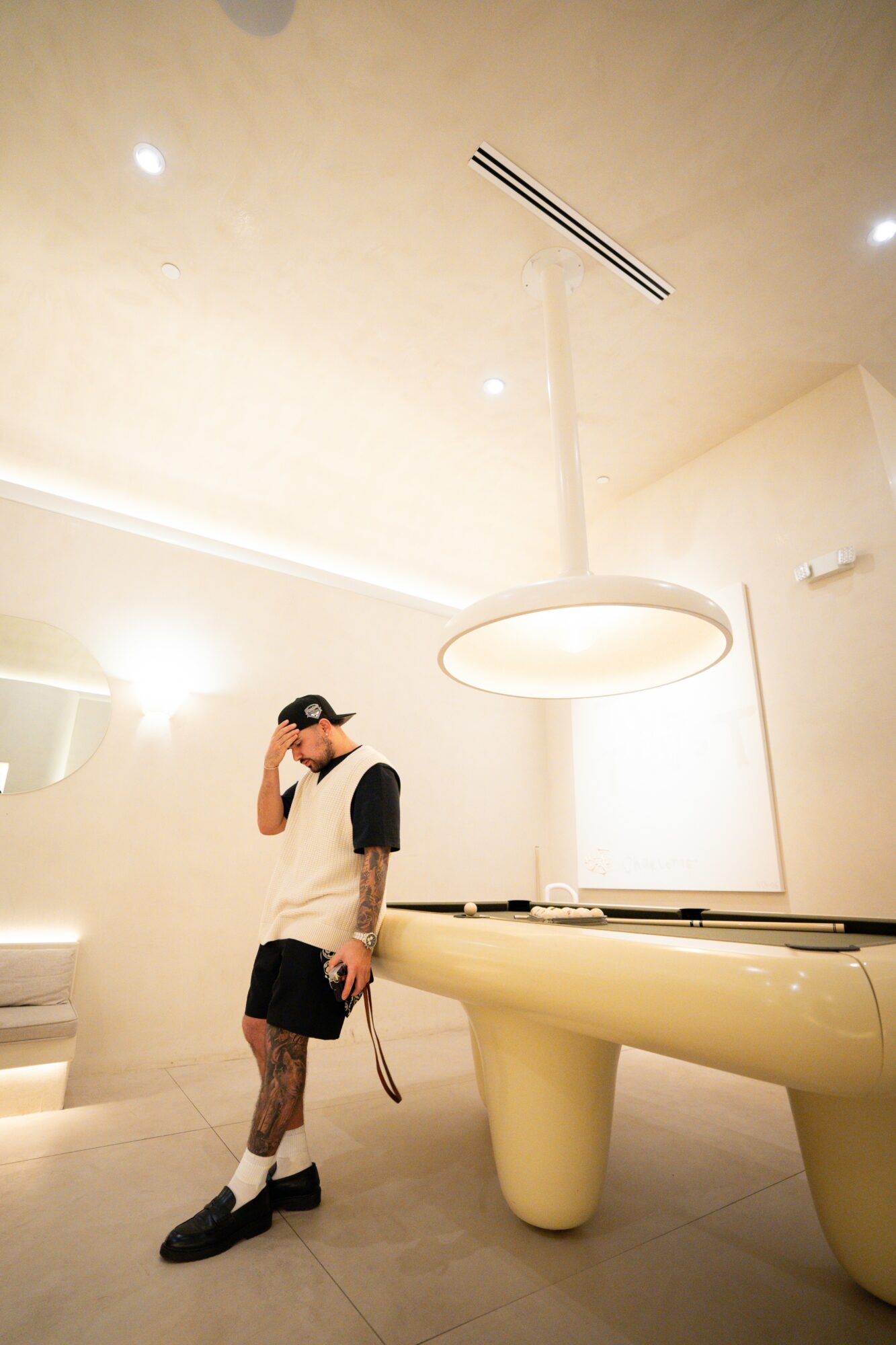 Person standing in a modern room with a large circular light fixture and a white table, holding a phone to their ear.