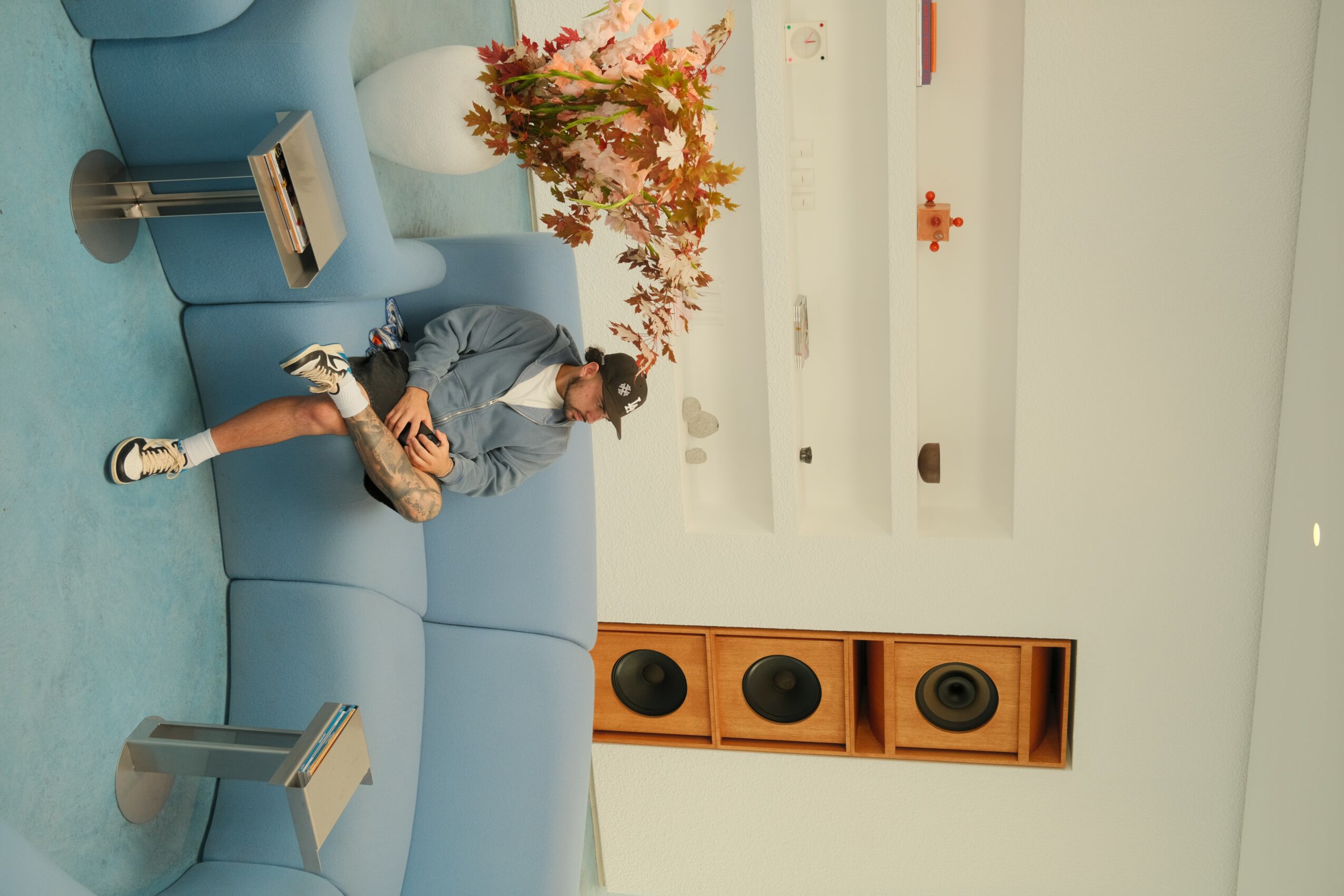 Person sitting on a blue sofa with a flower arrangement, in a bright room with white walls and wooden speaker unit.