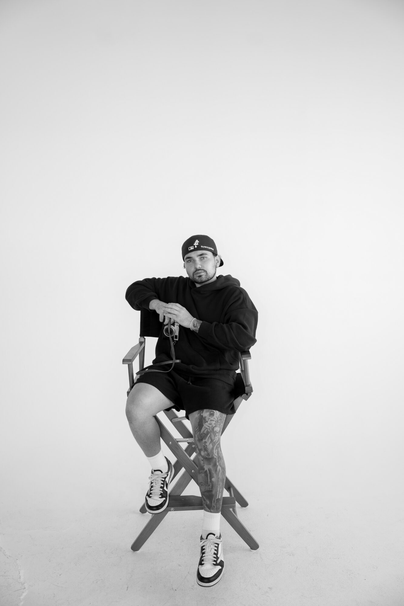 Person sitting on a chair with crossed legs, wearing a hoodie, shorts, sneakers, and a cap, against a plain background.