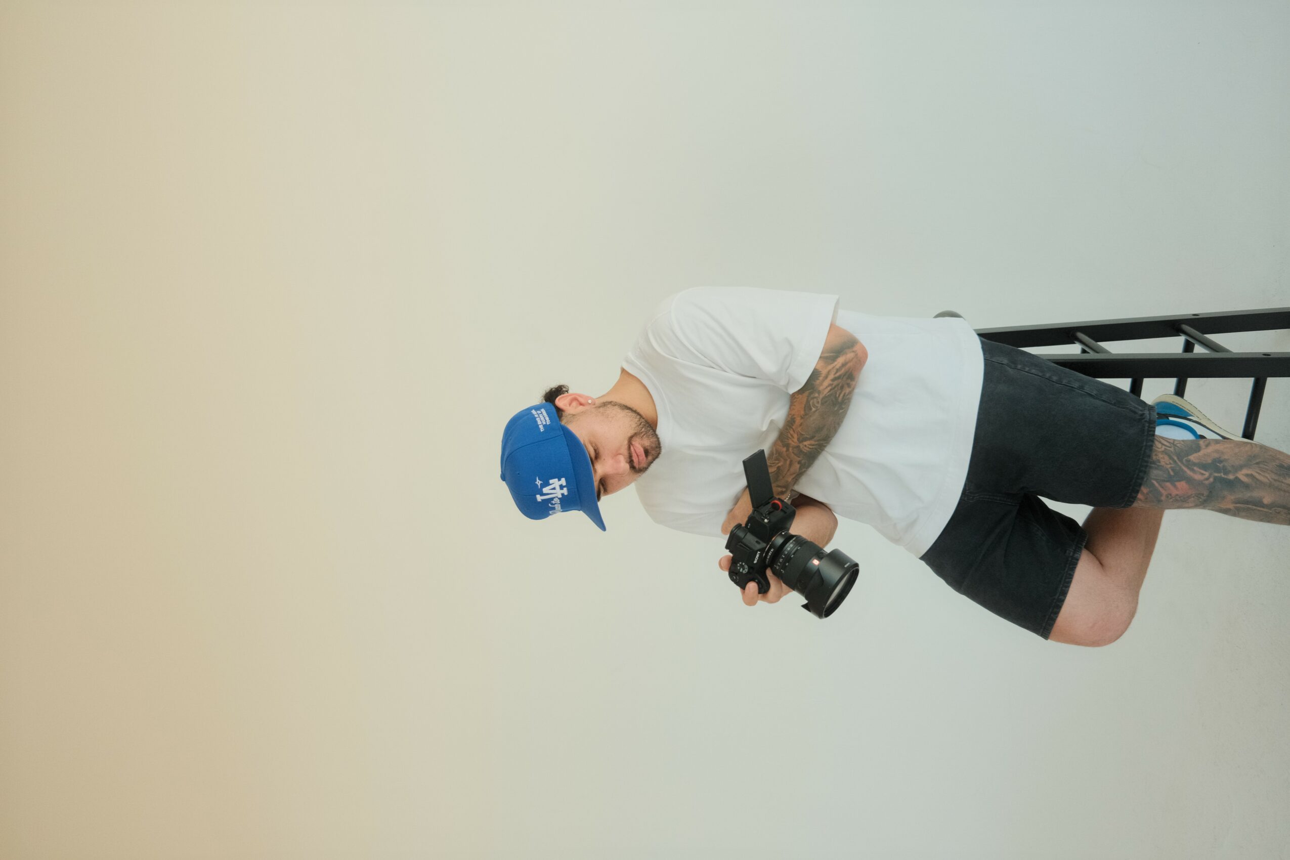 Person with tattoos holding a camera, wearing a white shirt, black shorts, and a blue cap, standing sideways.
