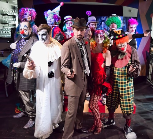 Group of people dressed as clowns and a man in a suit with a hat, posing on stage with colorful costumes.